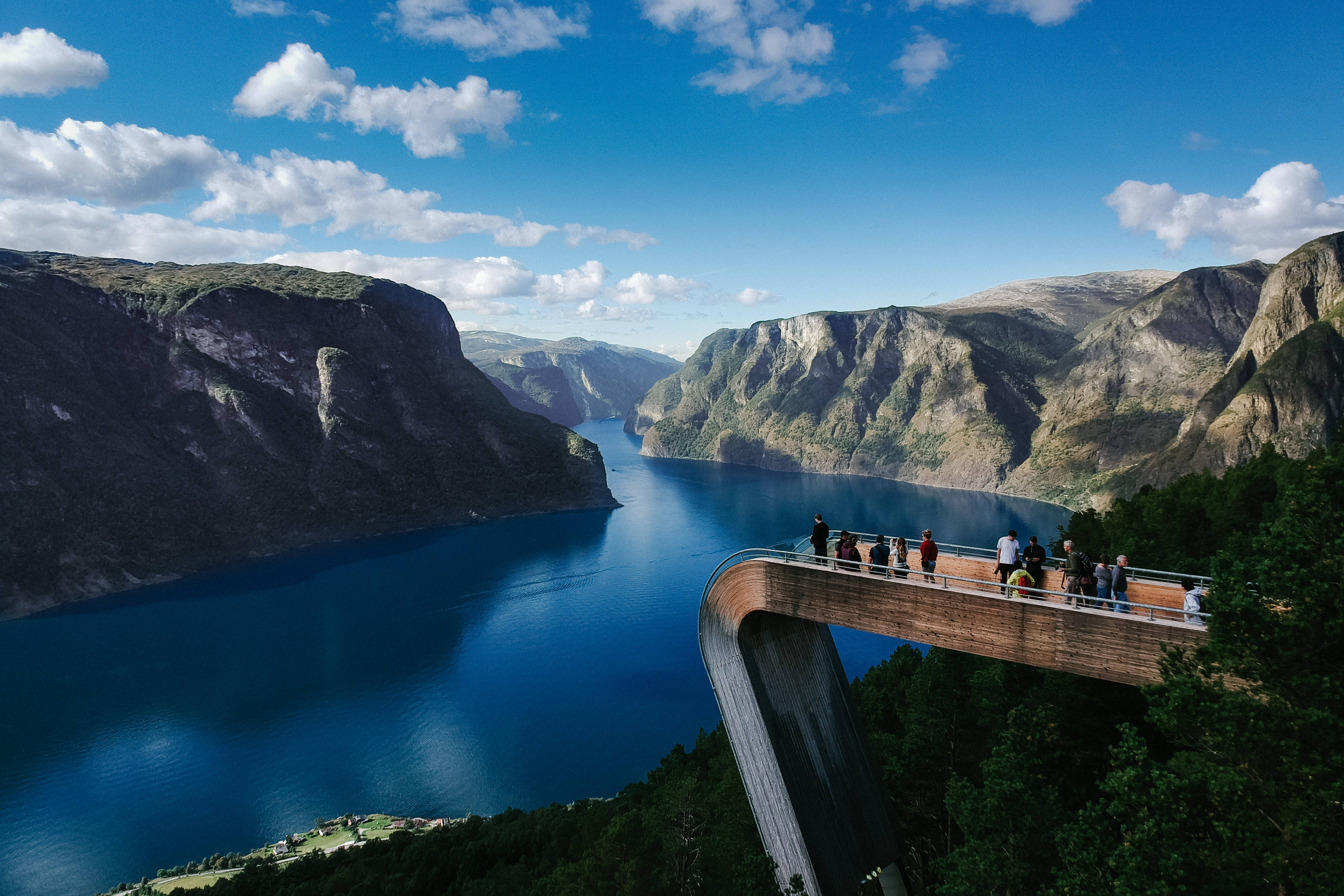 Norway 桌布 壁紙 — 遊客站在現代木質觀景台上，俯瞰深藍色峽灣，兩側是高聳的綠色山脈，頭頂是明亮的藍天和蓬鬆的白雲。