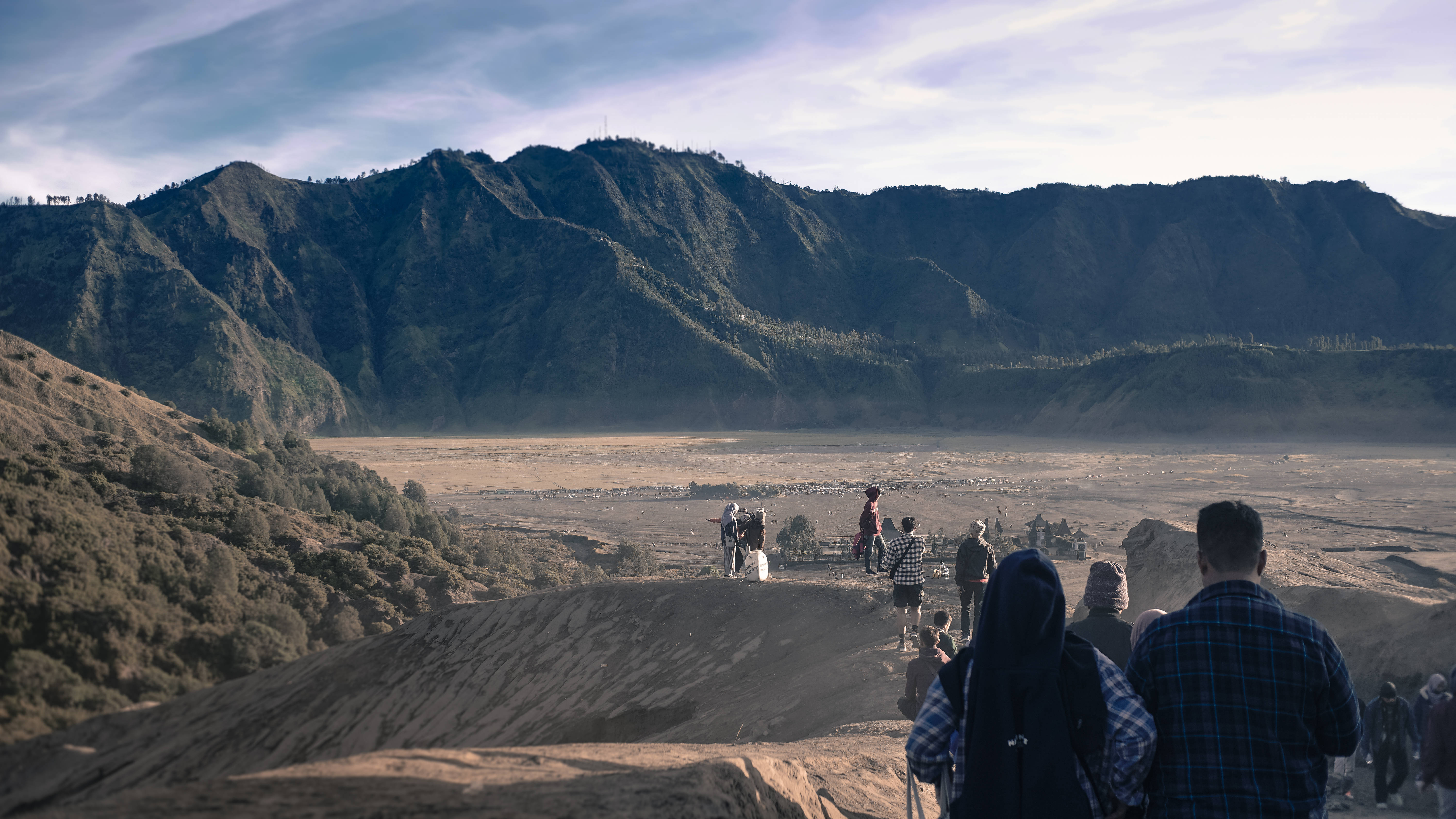 pemandangan 桌布 壁紙 — 遊客站在陽光普照的山脊上，俯瞰廣闊的火山口，森林覆蓋的山脈在部分多雲的天空下。