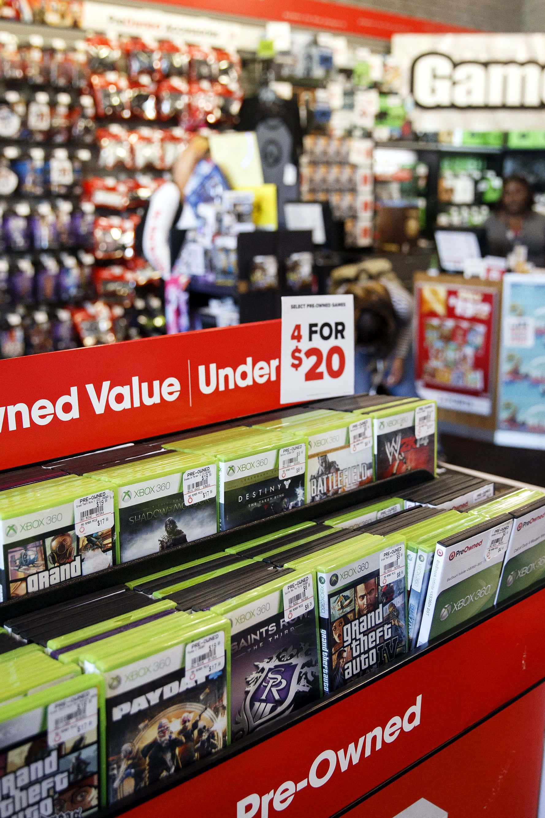 GameStop 桌布 壁紙 — 遊戲店內紅色二手貨架上Xbox 360遊戲盒的特寫，背景購物者模糊。