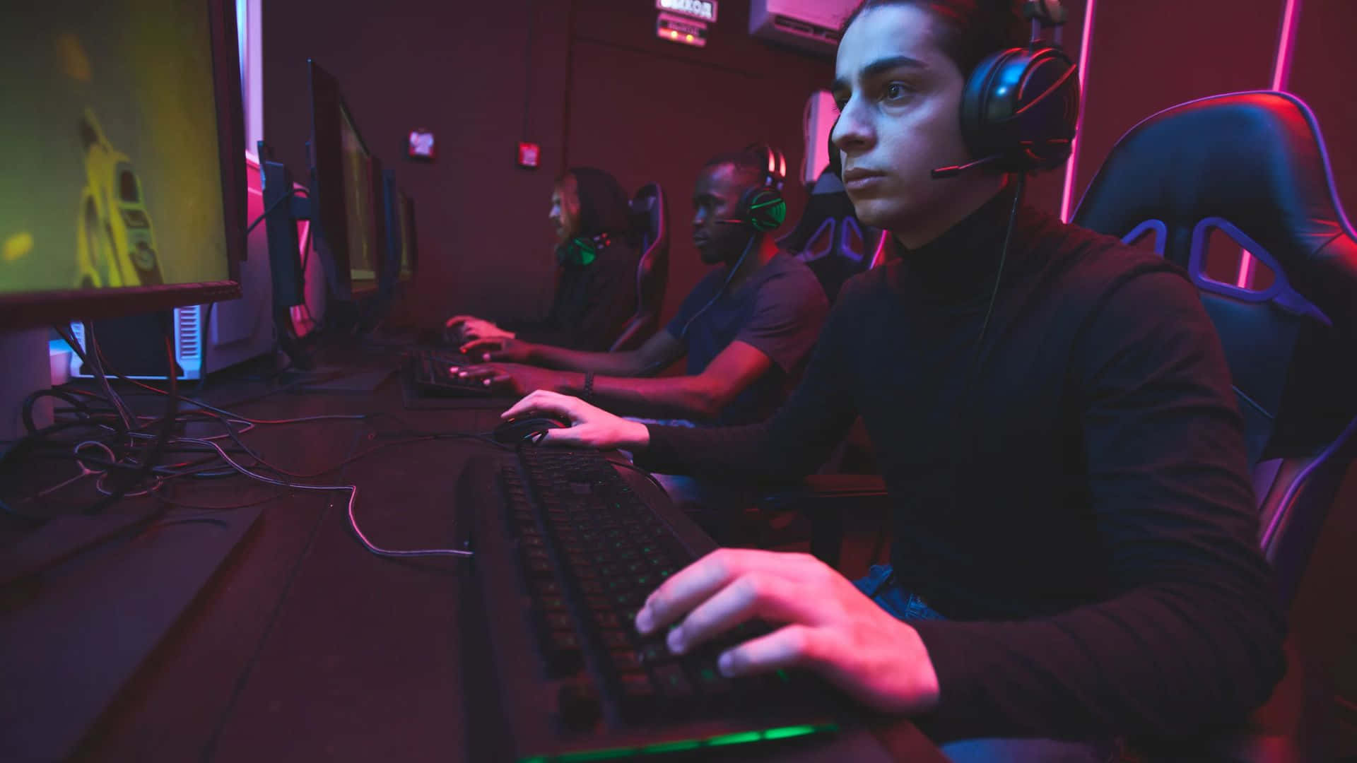 Gaming Tournaments 桌布 壁紙 — 遊戲玩家在昏暗的房間中，被霓虹粉紅和綠色燈光包圍，專注於發光的螢幕。