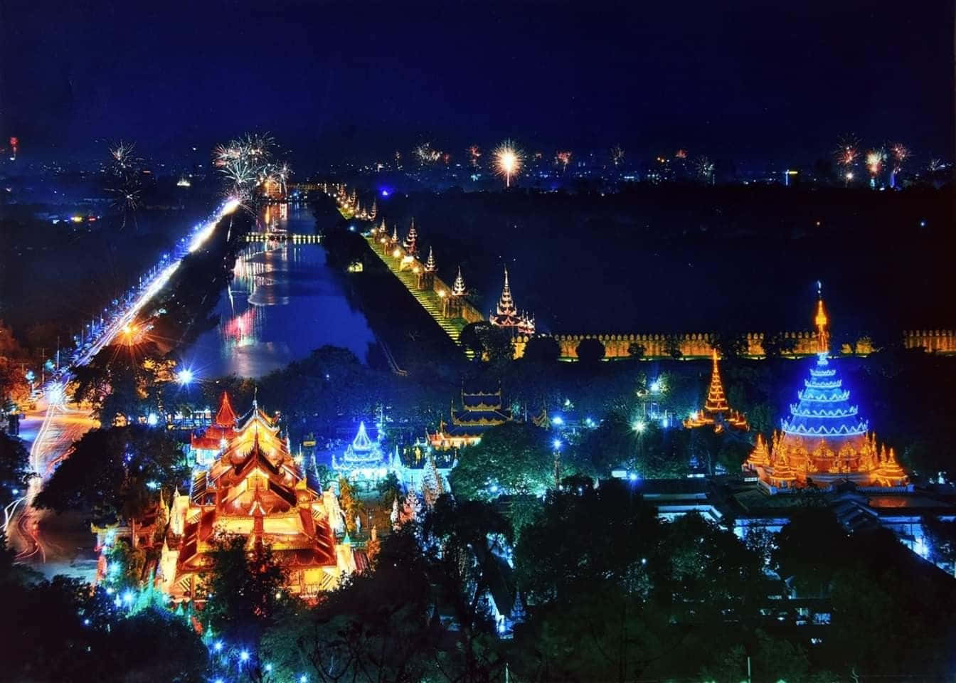 Light 桌布 壁紙 — 運河沿岸寺廟建築群的耀眼夜景，以鮮豔的藍色和金色燈光點亮，遠處有煙火綻放。
