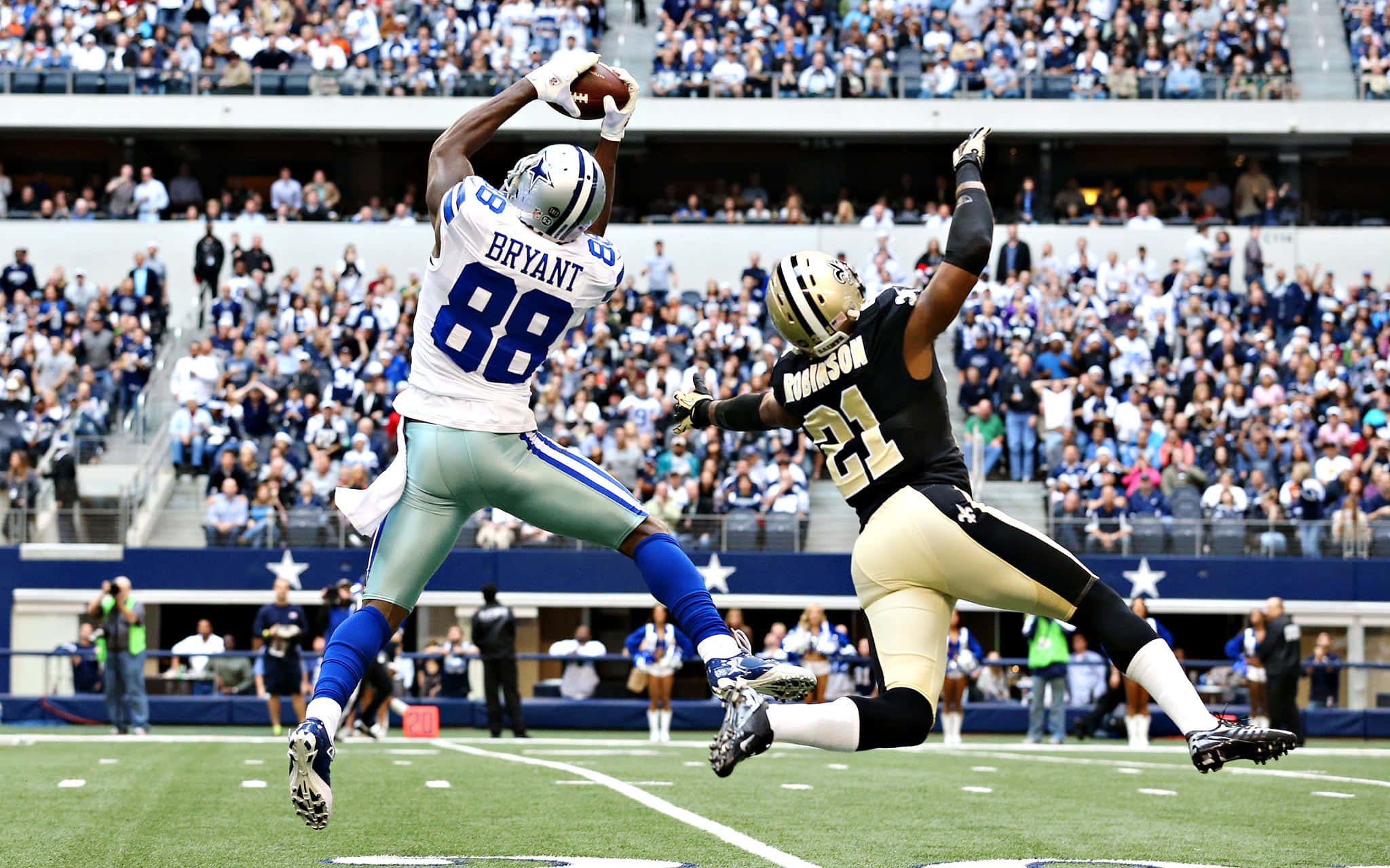 Dallas Cowboys Players 桌布 壁紙 — 達拉斯牛仔隊的外接手布萊恩特跳起接住橄欖球，而新奧爾良聖徒隊的防守球員羅賓遜則潛水試圖攔截，球場座無虛席。