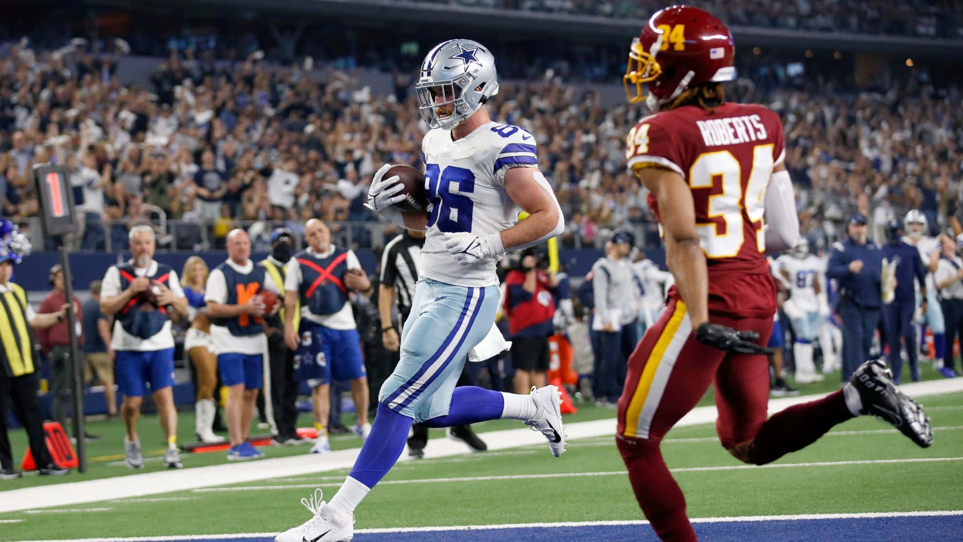 NFL 桌布 壁紙 — 達拉斯牛仔隊的外接手身穿白色球衣，帶著橄欖球奔跑，而華盛頓隊的紅色防守球員在燈火通明的球場上追趕他。