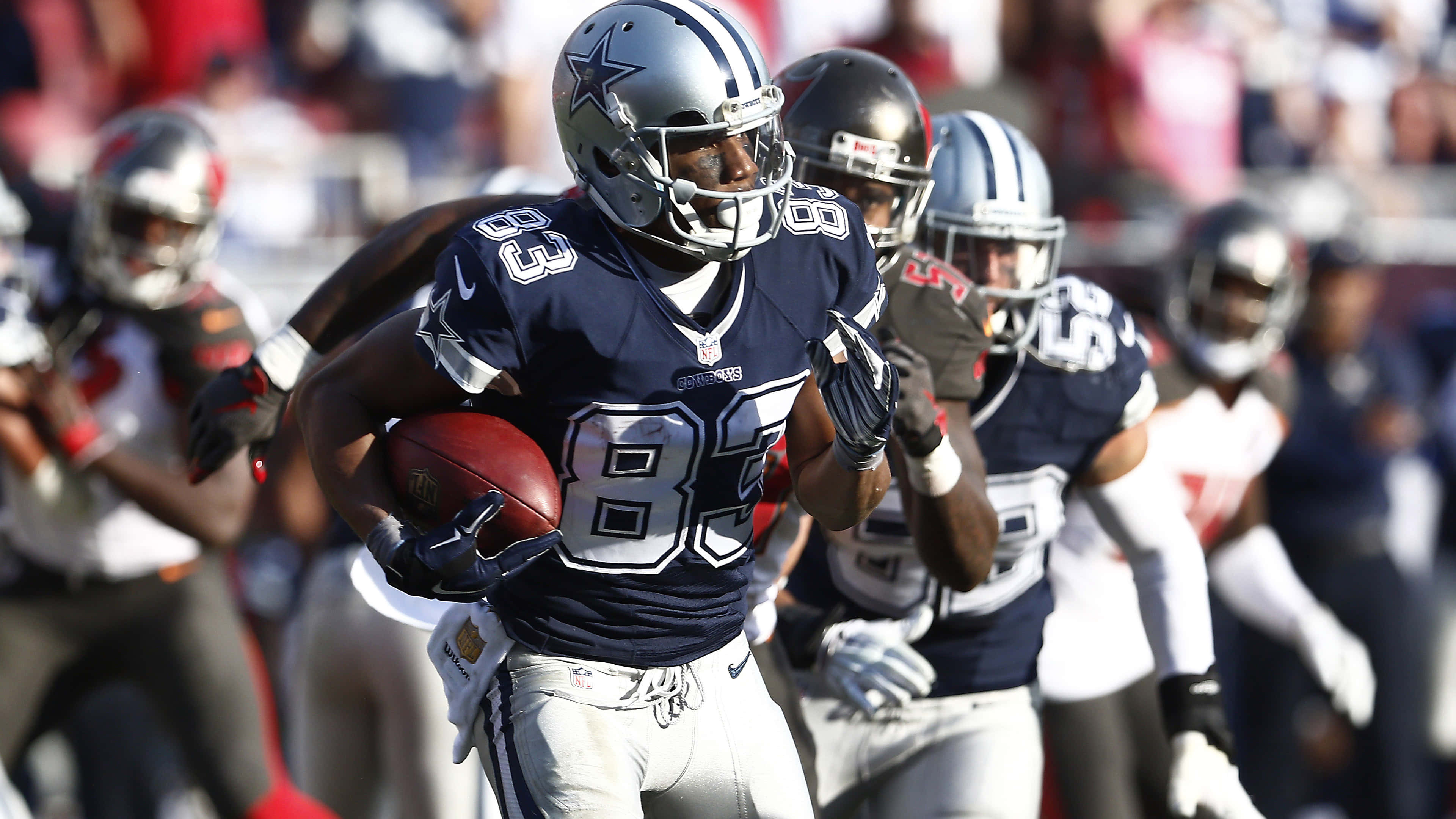 Dallas Cowboys Players 桌布 壁紙 — 達拉斯牛仔隊的外接手身穿藍銀色制服，在日間比賽中帶著橄欖球奔跑，背景是模糊的隊友和人群。
