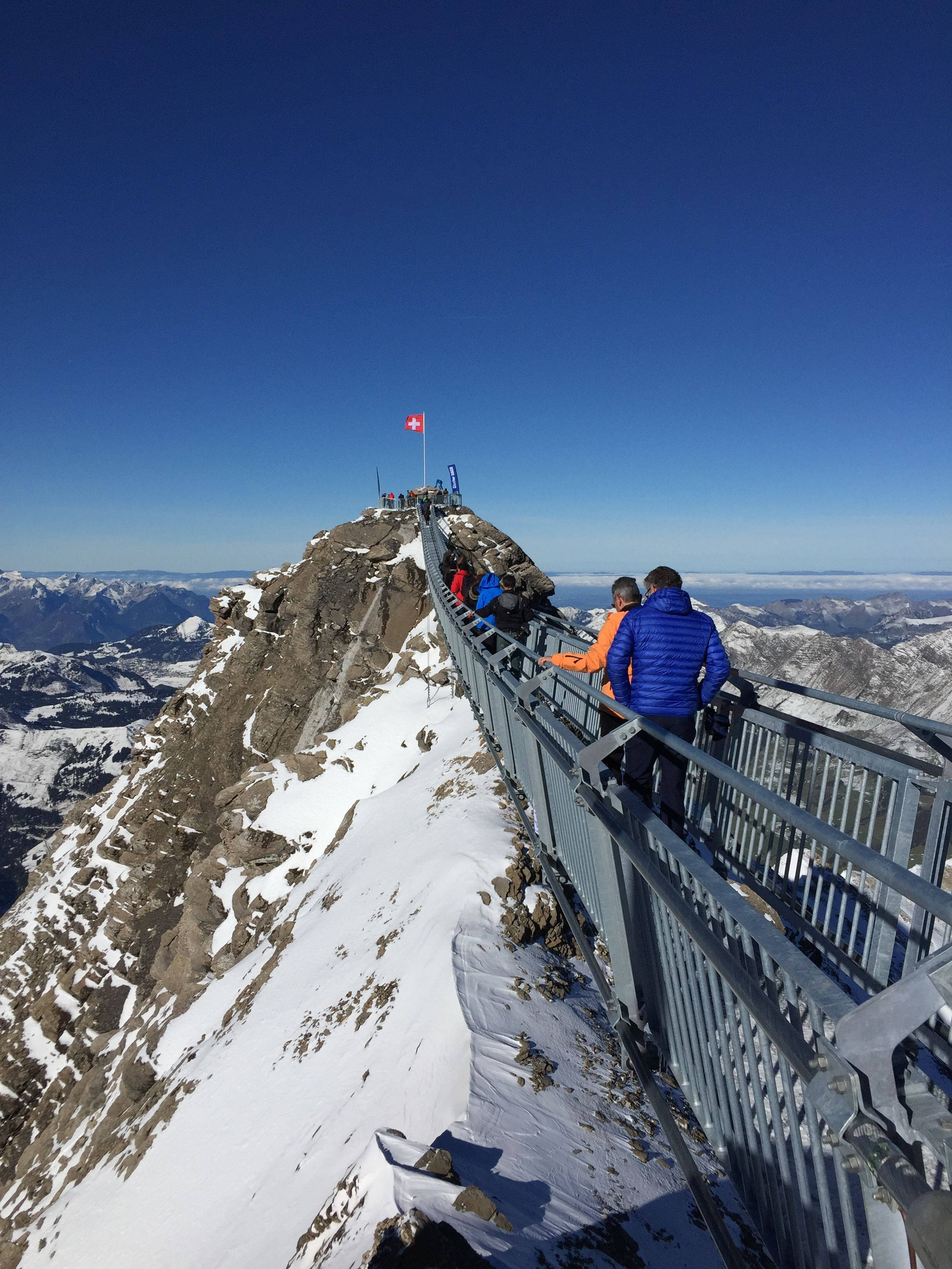 Alps 桌布 壁紙 — 遠足人士沿著一條狹窄的金屬橋行走，橋樑緊貼著積雪的山脊，天空清澈湛藍，山頂上飄揚著瑞士國旗。