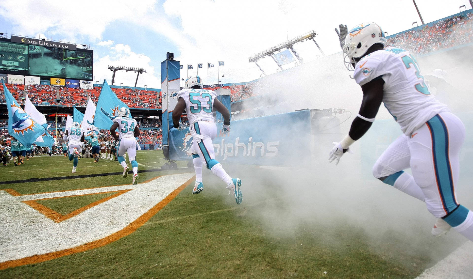 dolphins 桌布 壁紙 — 邁阿密海豚隊美式足球員在日間比賽中衝過煙霧隧道進入太陽生活體育場的球場。
