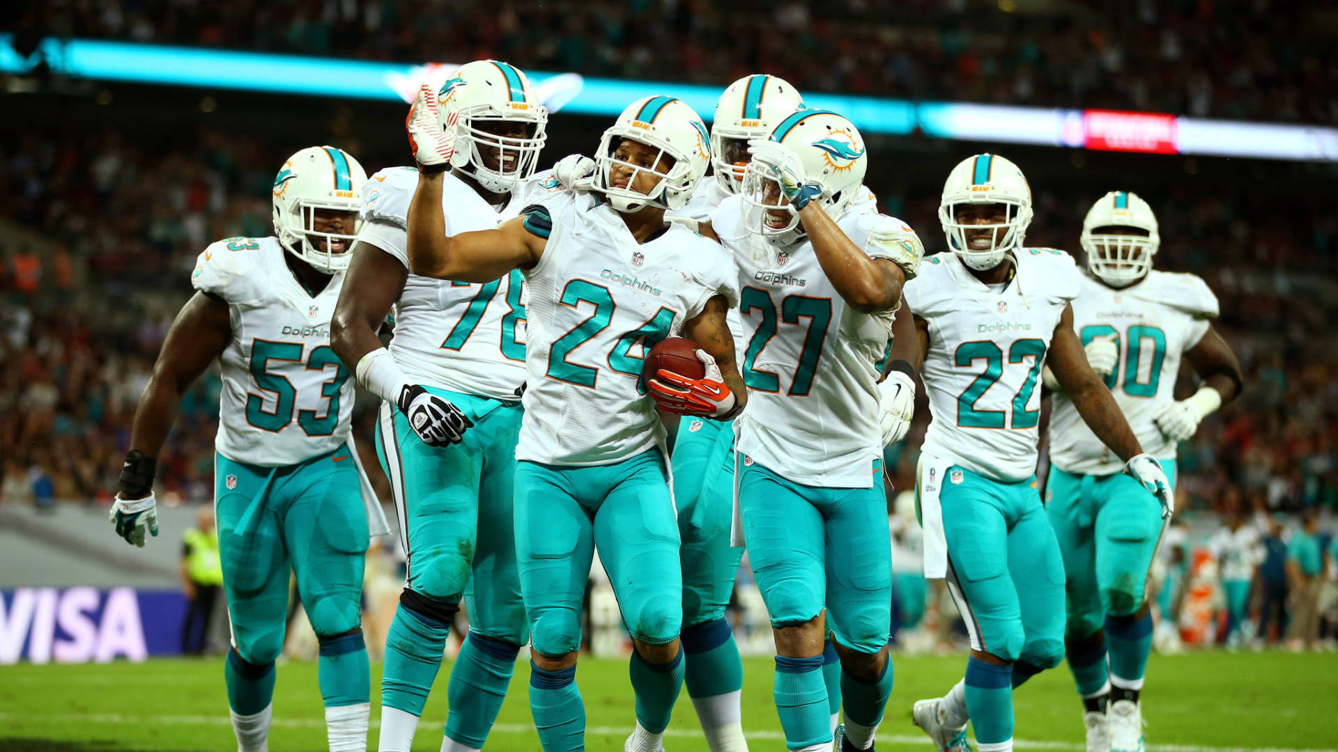 dolphins 桌布 壁紙 — 邁阿密海豚隊美式足球員身穿白色和藍綠色球衣，在燈光明亮的球場上慶祝達陣。