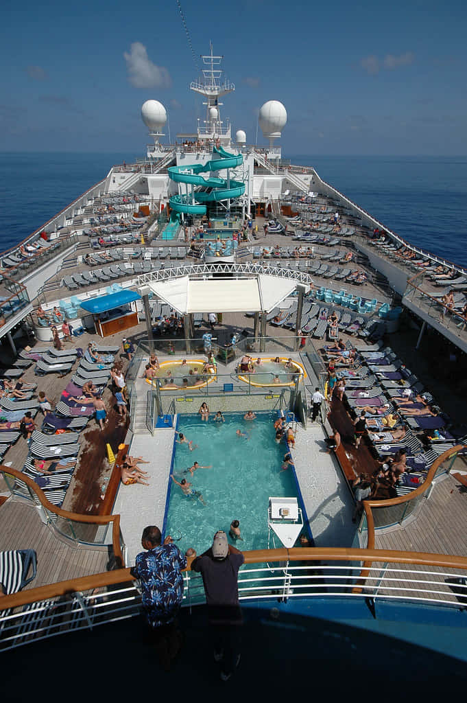 Carnival Conquest 桌布 壁紙 — 郵輪甲板的鳥瞰圖，在晴朗的藍天下，甲板上擠滿了在泳池和滑梯旁享受日光浴的人們。