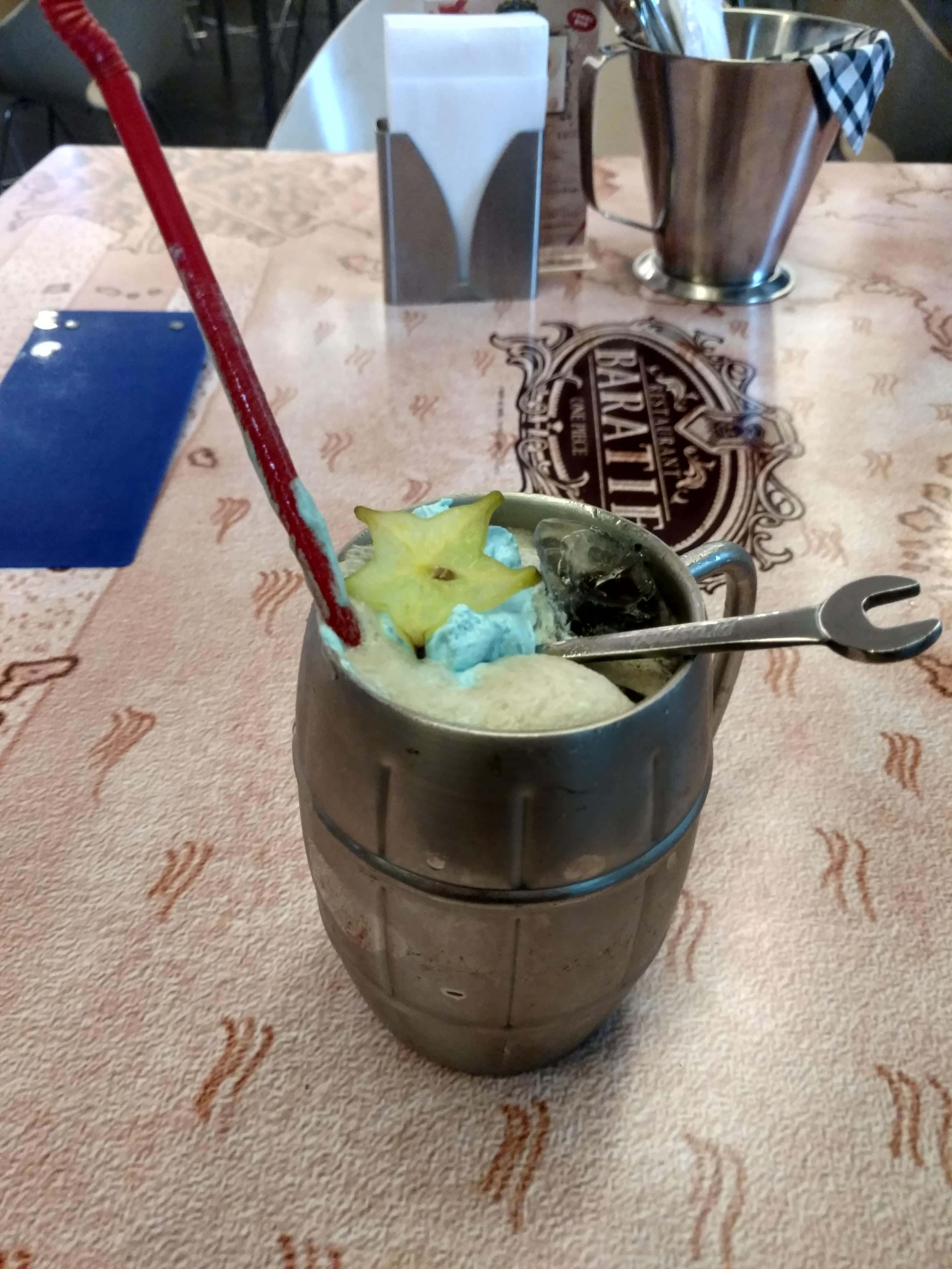 Baratie 桌布 壁紙 — 酒吧內，一個金屬杯盛裝著冰凍藍色飲料，上面放有楊桃和紅色吸管，放在圖案桌巾上。