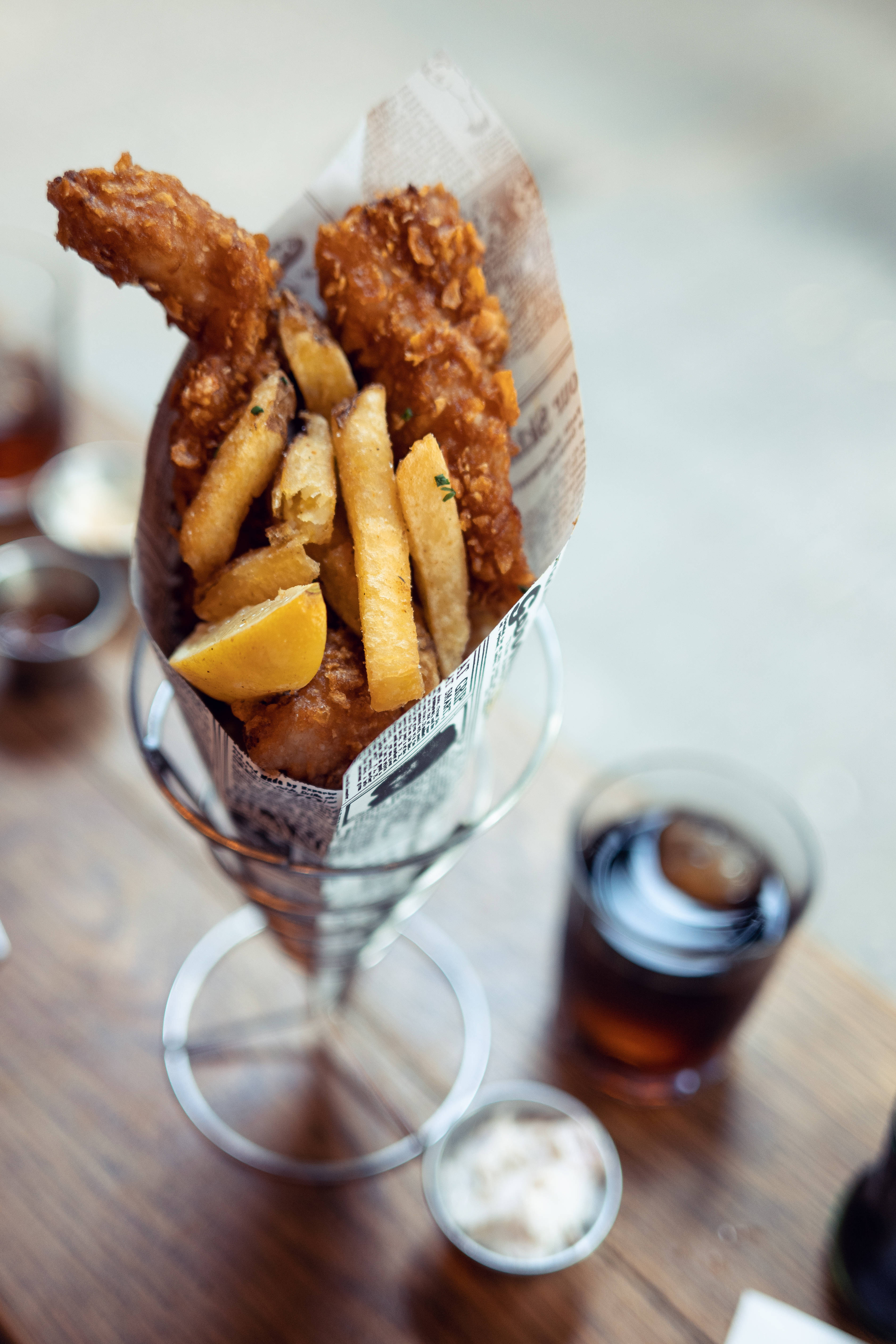 魚 桌布 壁紙 — 酥脆炸雞和金黃薯條包裹在報紙中，置於玻璃錐體內，放在木桌上，附有蘸醬和汽水。