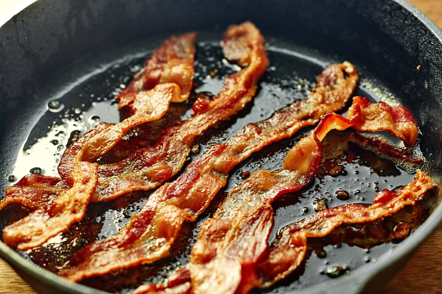 bacon 桌布 壁紙 — 酥脆的培根條在黑色鑄鐵煎鍋中滋滋作響，帶有冒泡的油脂和木紋背景。