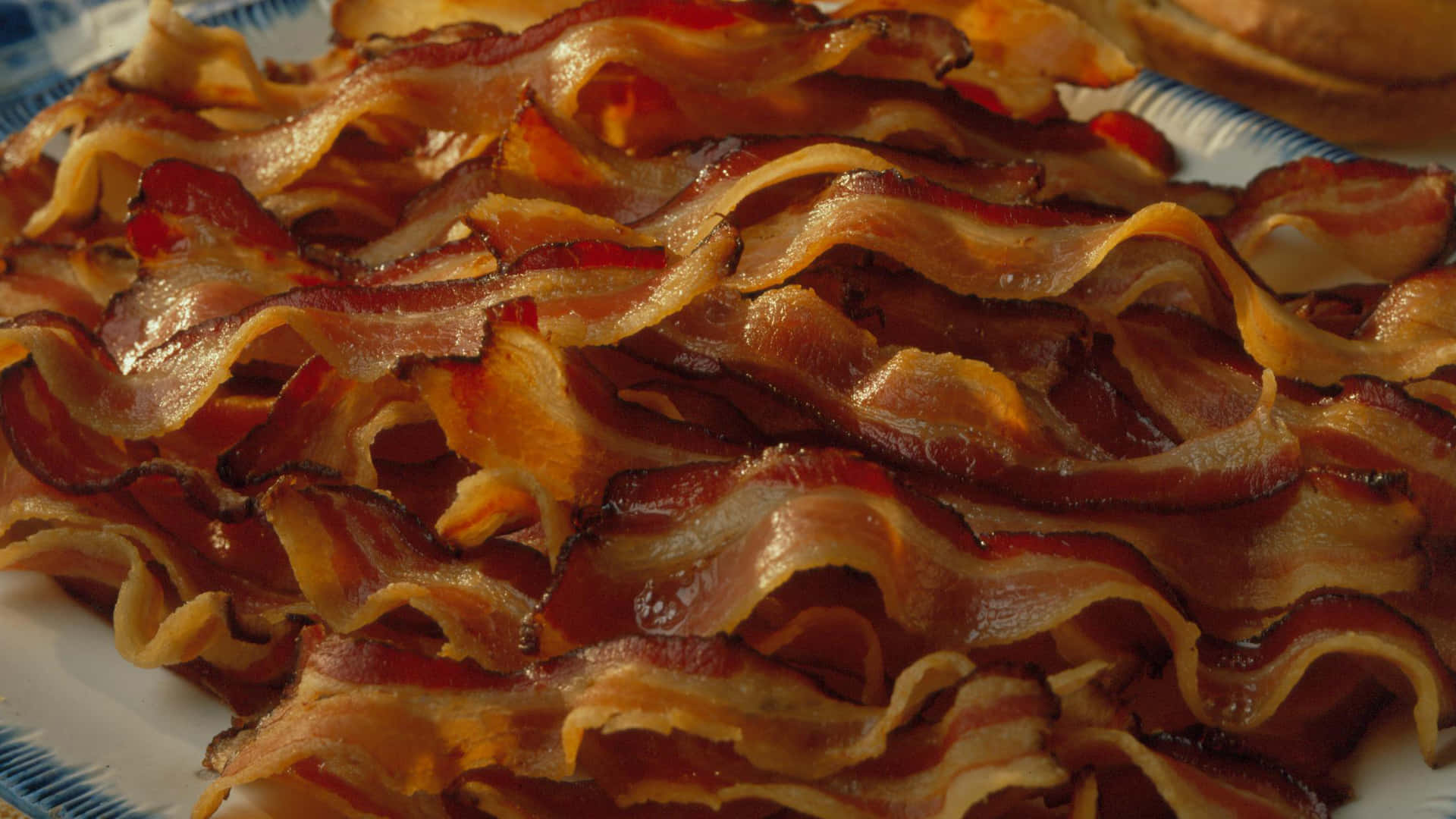 bacon 桌布 壁紙 — 酥脆金黃的培根條閃爍著油脂，在帶有圖案的盤子上誘人地捲曲。