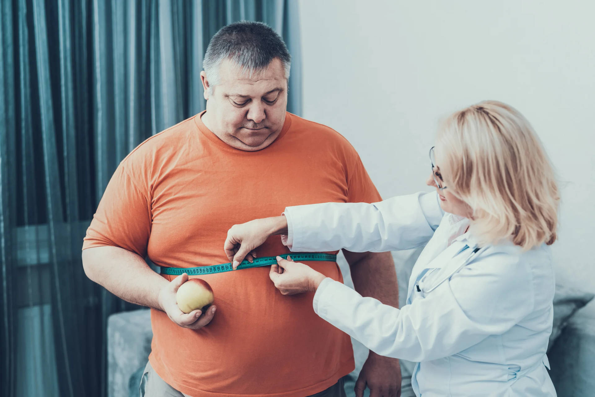 Health 桌布 壁紙 — 醫生用軟尺量測一名男性的腰圍，而該男性在臨床環境中手持一個蘋果。