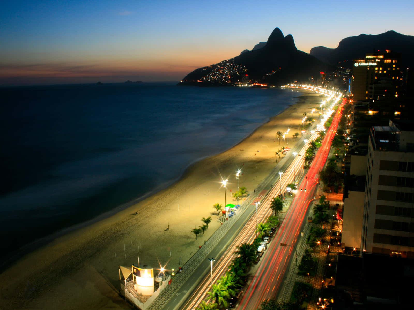 Brazilian Beach 桌布 壁紙 — 里約海濱充滿活力的黃昏景色，發光的街燈和流動的車燈映襯著漸暗的海洋和標誌性的山脈。