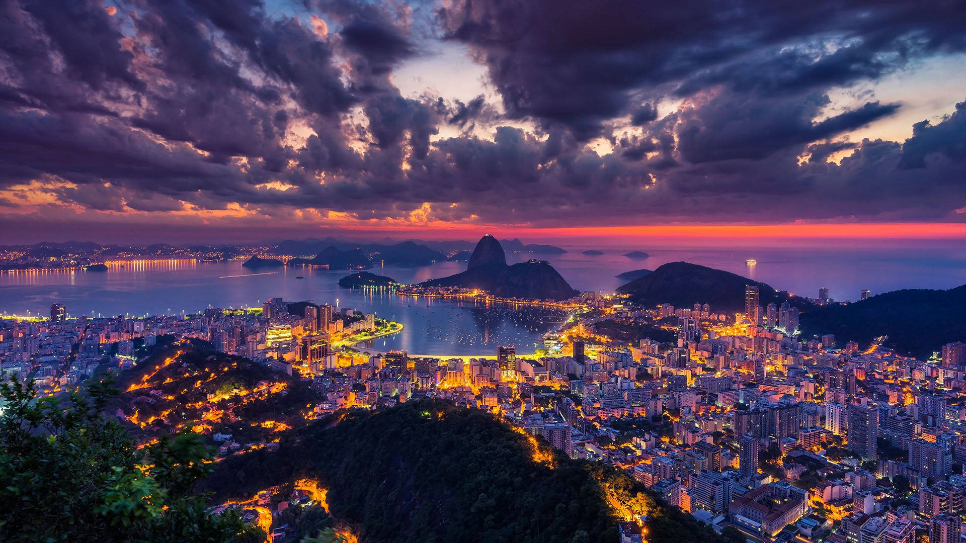 South America 桌布 壁紙 — 里約熱內盧令人驚嘆的日落，甜麵包山和城市燈光在戲劇性的紫色和橙色雲層下閃耀。