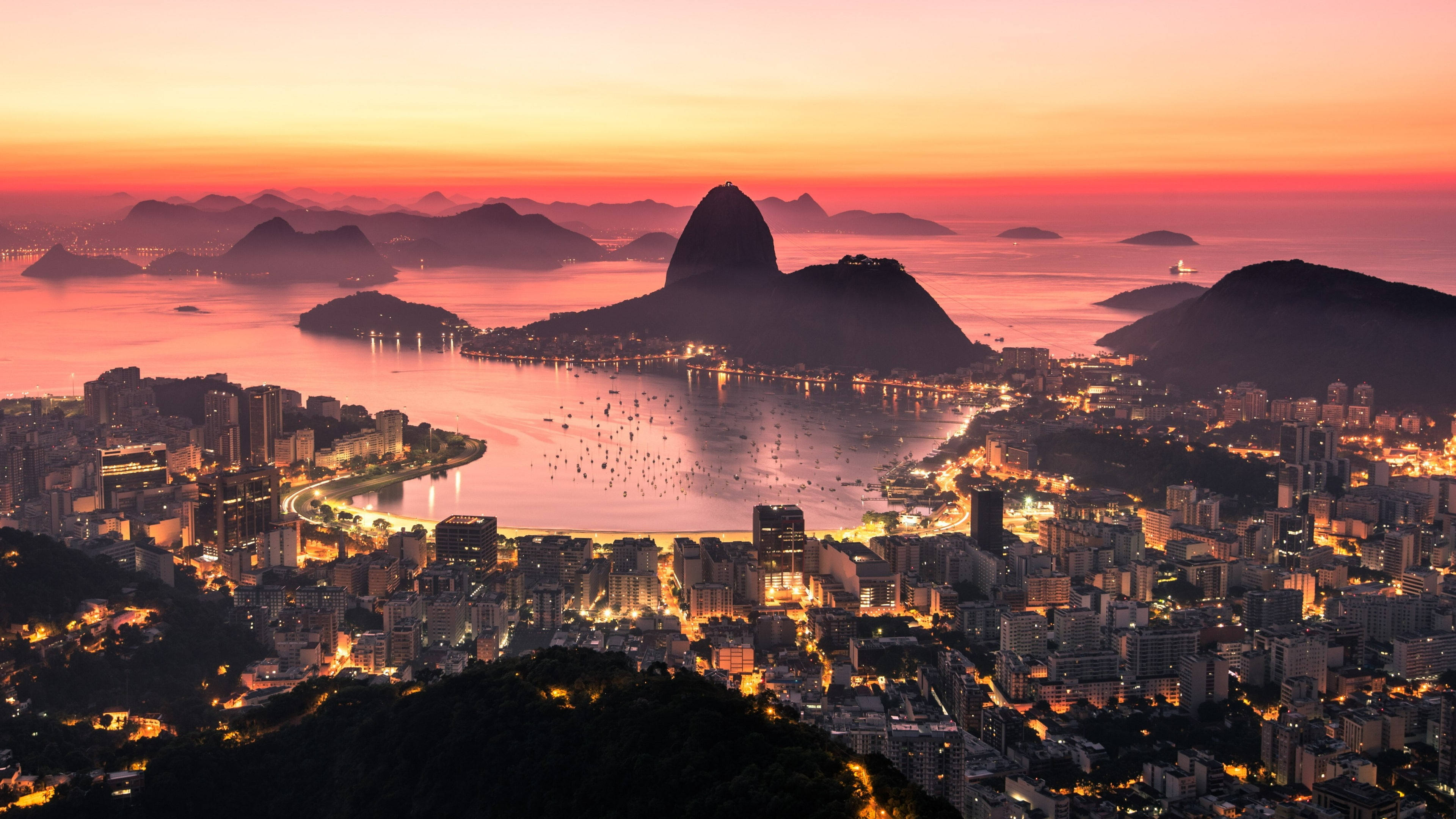rio 桌布 壁紙 — 里約熱內盧令人驚嘆的日落，甜麵包山在熾熱的橙色和粉紅色天空下形成剪影，城市燈光在下方閃爍。