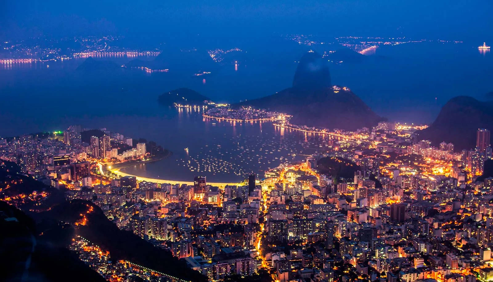 South America 桌布 壁紙 — 里約熱內盧夜景鳥瞰圖，城市景觀在群山與海洋之間閃耀，標誌性的甜麵包山亮起燈光。