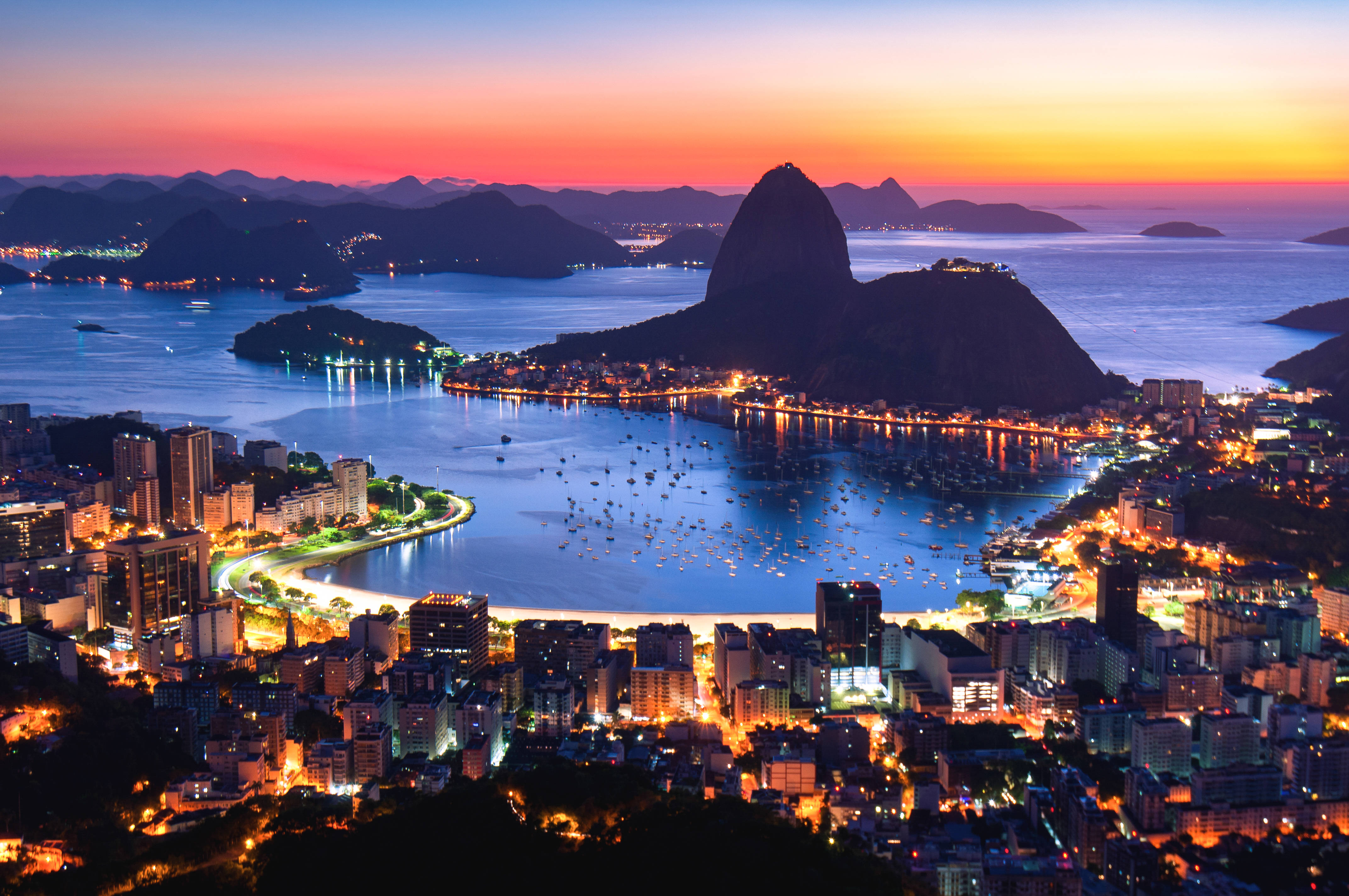 South America 桌布 壁紙 — 里約熱內盧日落時分，甜麵包山的剪影映襯著火紅橙粉色的天空，下方是閃爍的城市燈光與水面。