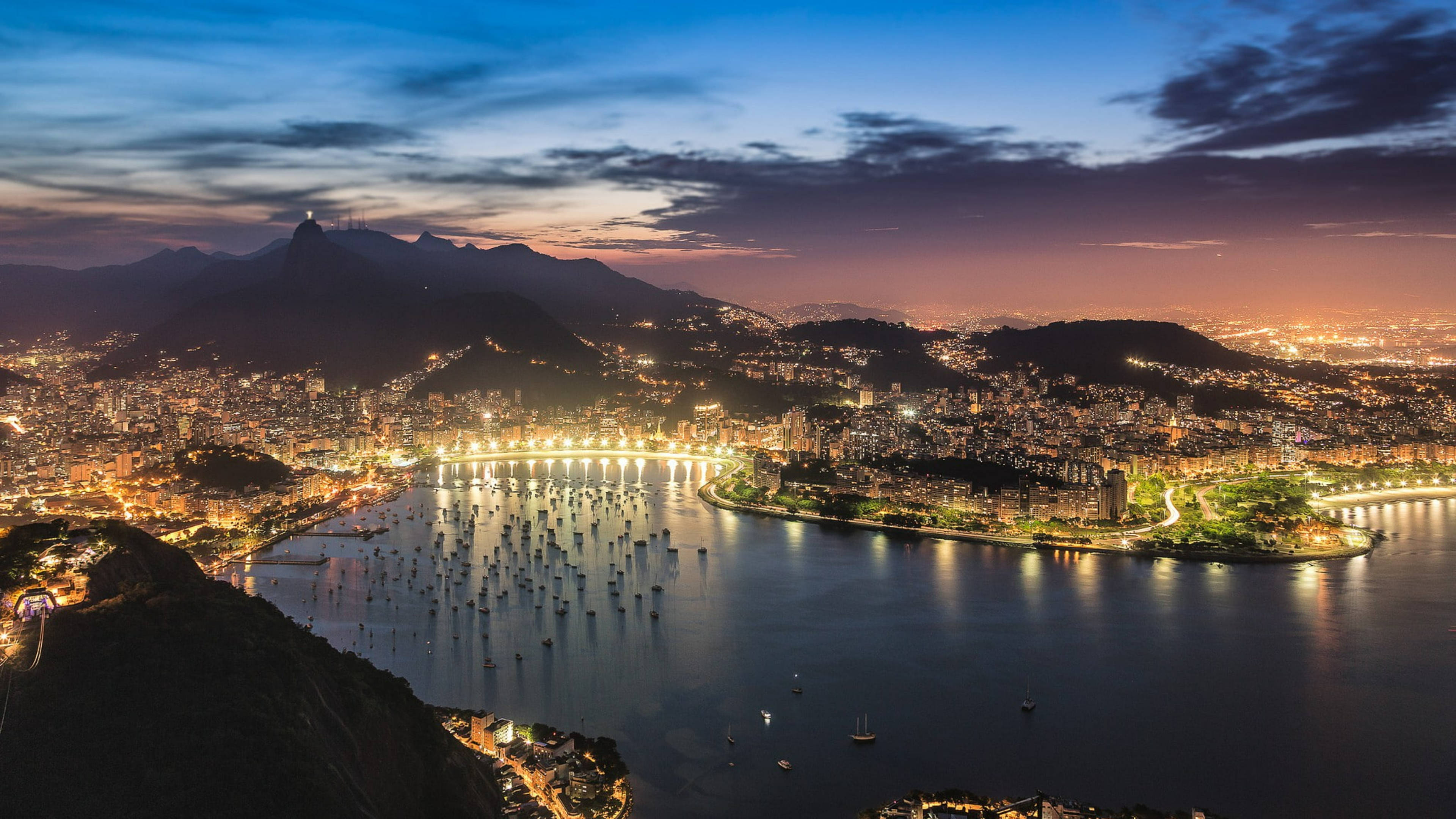 rio 桌布 壁紙 — 里約熱內盧璀璨的城市景觀沿著閃爍的海灣，背景是山脈，令人嘆為觀止的黃昏景色。