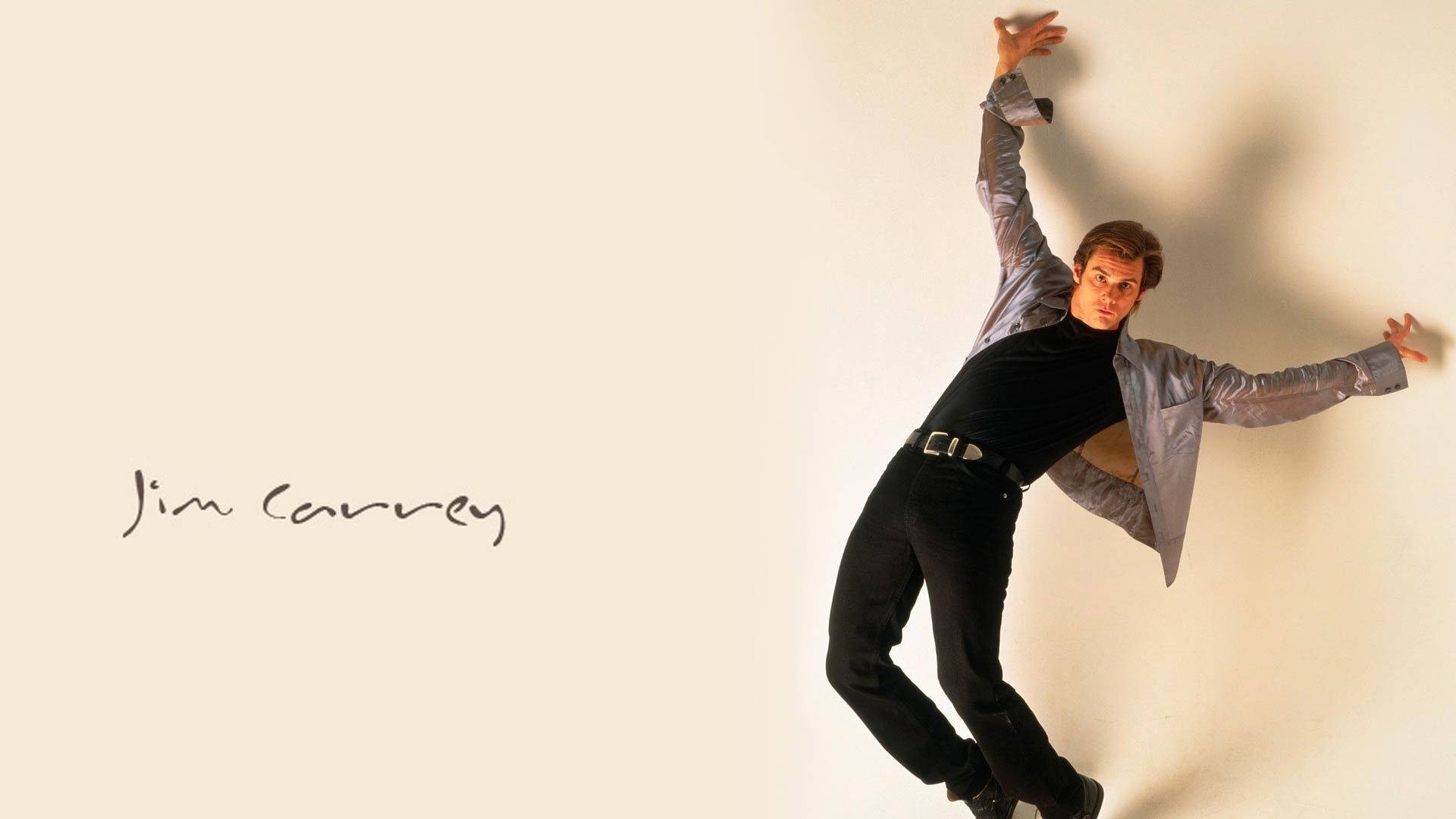 Jim Carrey 桌布 壁紙 — 金凱瑞穿著灰色外套和黑色褲子，以動態姿勢靠著素牆。