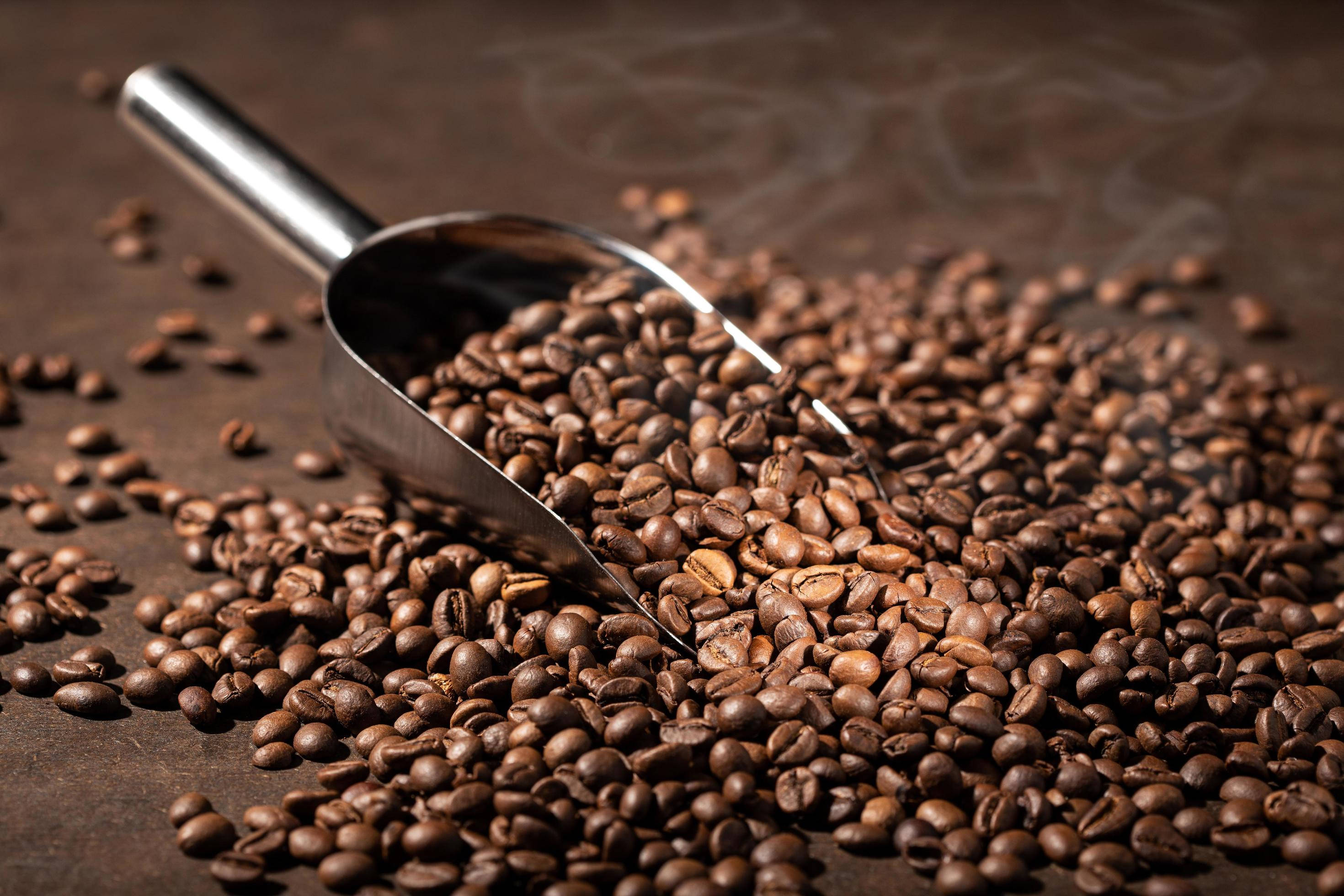 beans 桌布 壁紙 — 金屬勺子將濃郁的棕色烘焙咖啡豆灑落在深色鄉村風格的表面上，縷縷蒸汽升起。