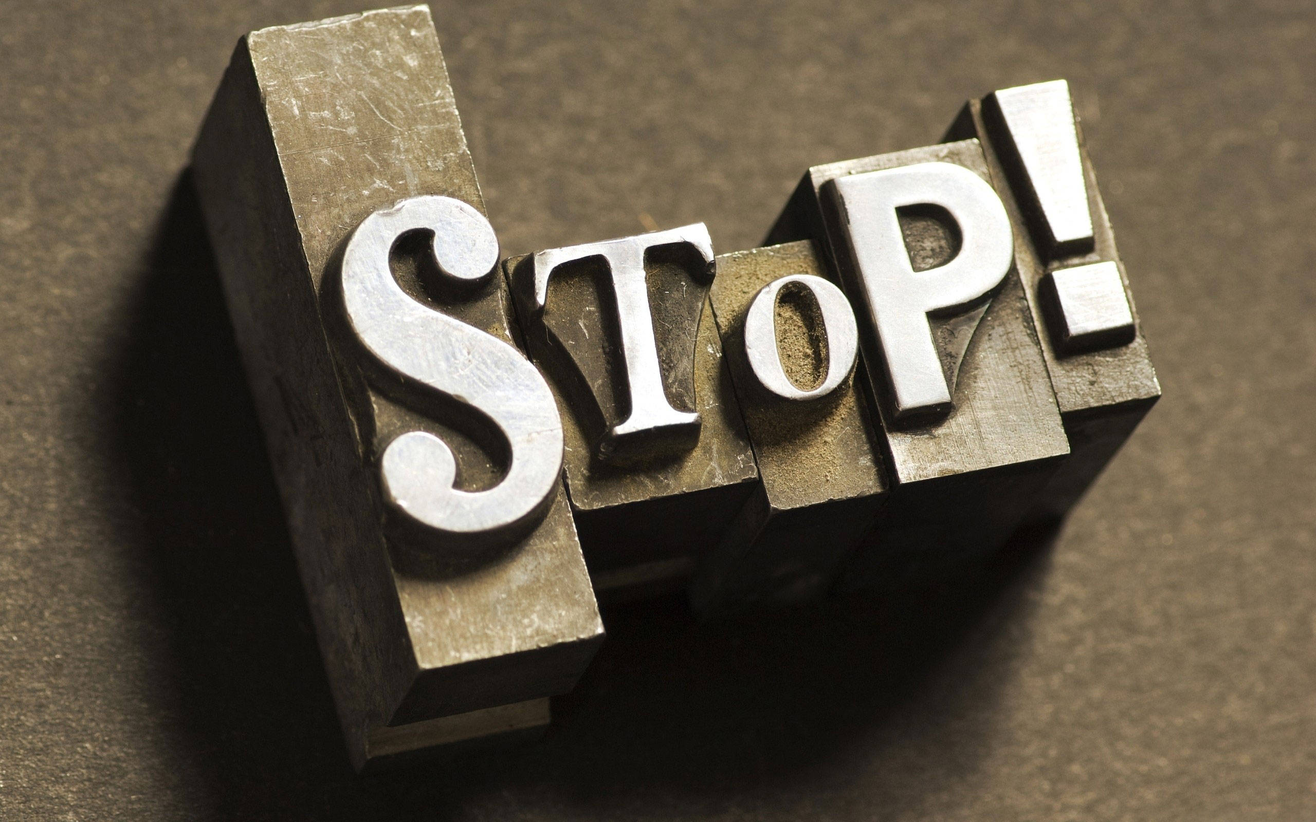 Typography 桌布 壁紙 — 金屬活字塊拼出「STOP!」，在戲劇性燈光下於有紋理的棕色表面投下銳利陰影。