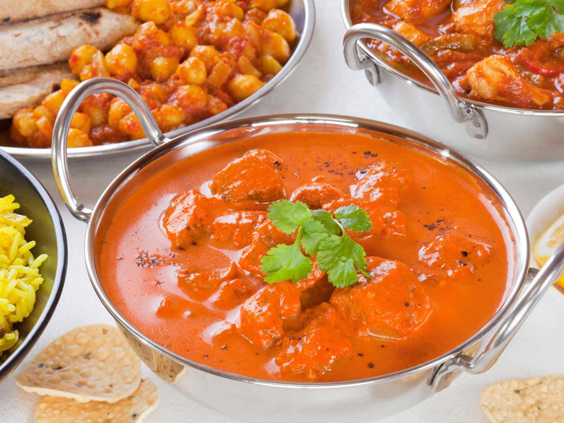 Indian 桌布 壁紙 — 金屬碗中的濃郁橙色咖哩，搭配雞肉塊和香菜裝飾，周圍環繞著印度烤餅和黃米飯。