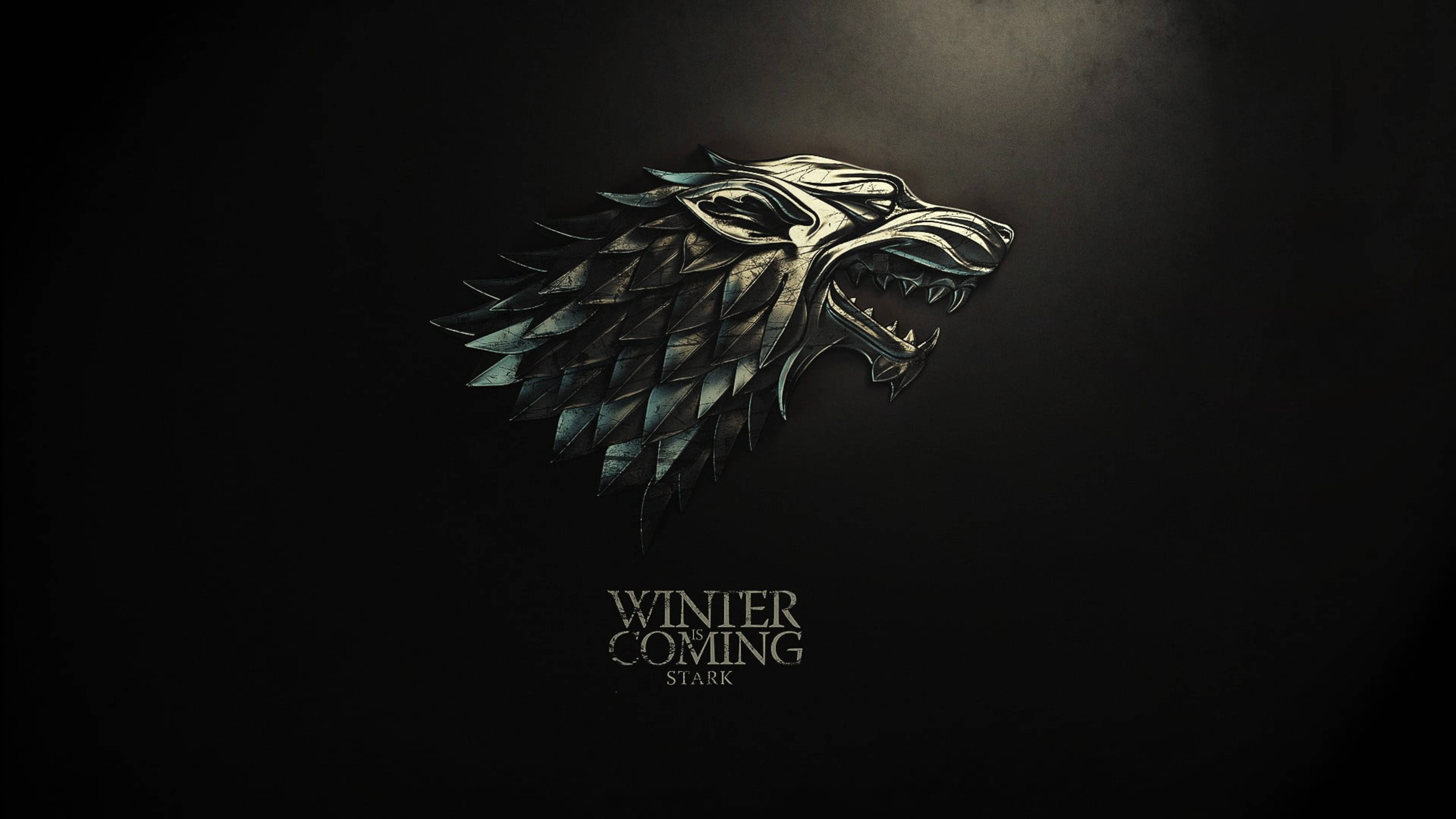 House Stark 桌布 壁紙 — 金屬製的冰原狼頭徽章在聚光燈下於深色背景上發光，並帶有「Winter is Coming Stark」文字。