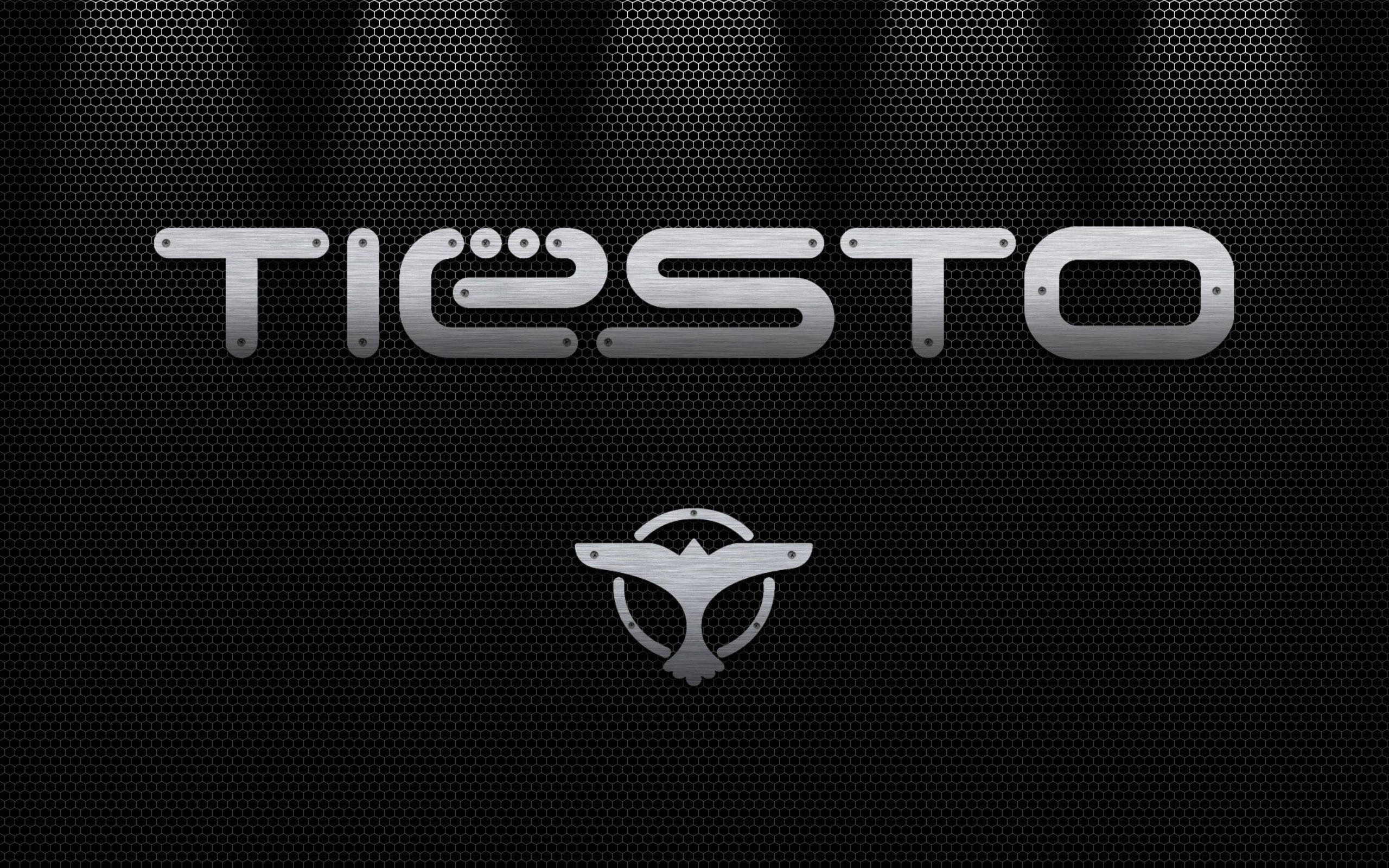 Tiesto 桌布 壁紙 — 金屬質感 'TIËSTO' 標誌，帶有翼形徽章，位於黑色穿孔背景上，聚光燈光束照射。