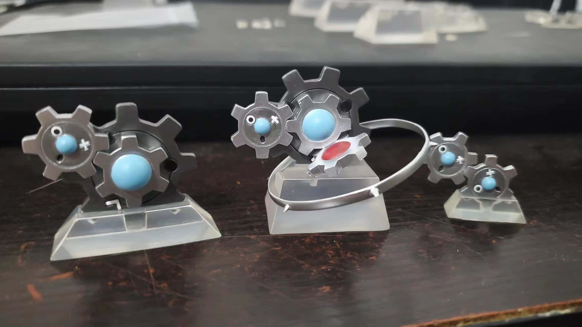 寶可夢 桌布 壁紙 — 金屬齒輪公仔，藍色眼睛和紅色點綴，排列在透明支架上，置於深色木質表面。