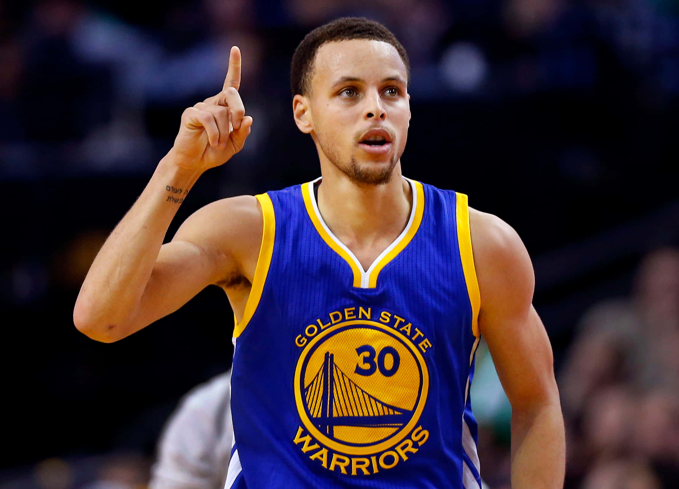 NBA 桌布 壁紙 — 金州勇士籃球運動員史蒂芬柯瑞身穿藍黃球衣在比賽中向上指。