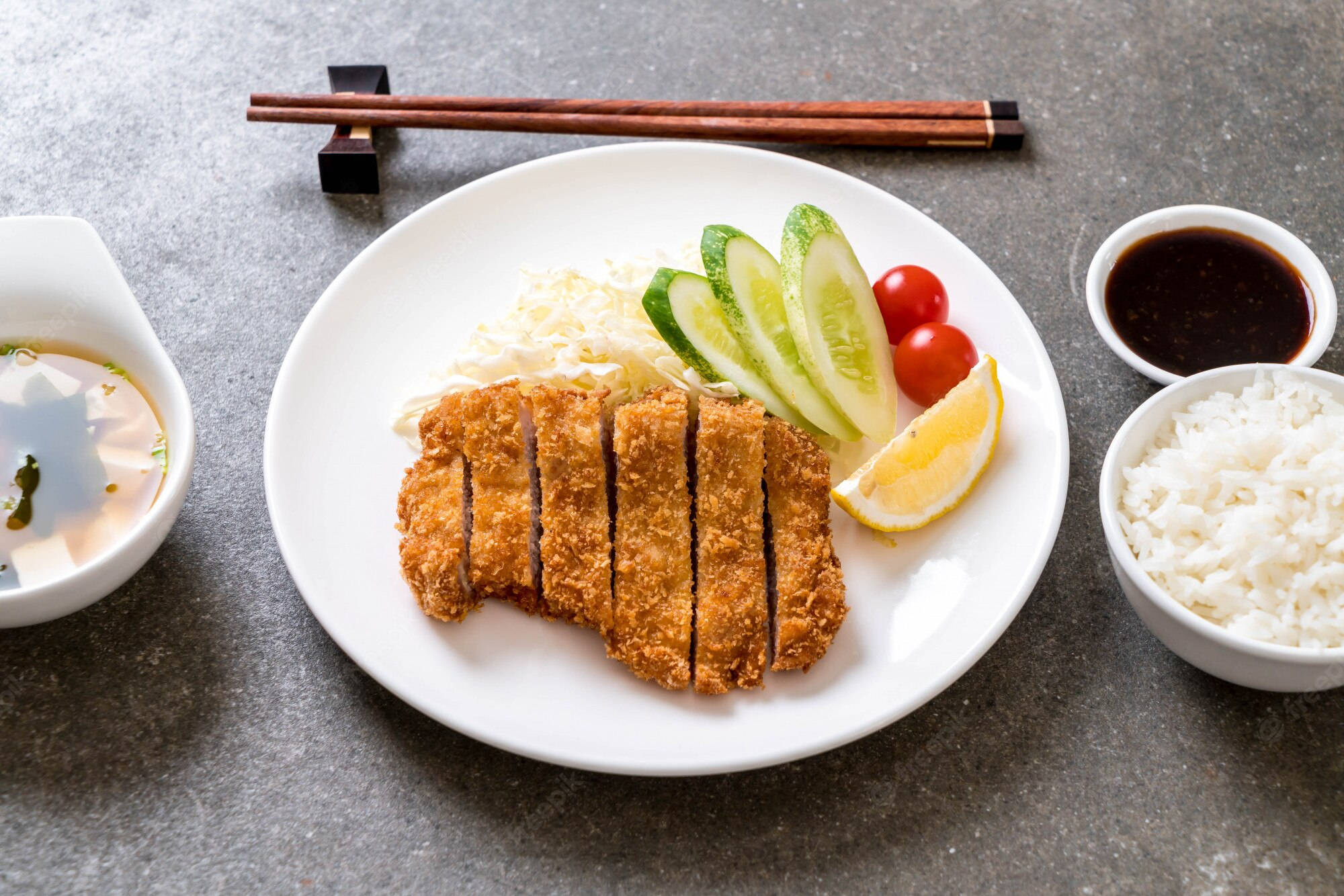 Food Photography 桌布 壁紙 — 金棕色切片炸豬排放在白色盤子上，搭配米飯、小黃瓜、番茄和醬汁，置於灰色石面上。