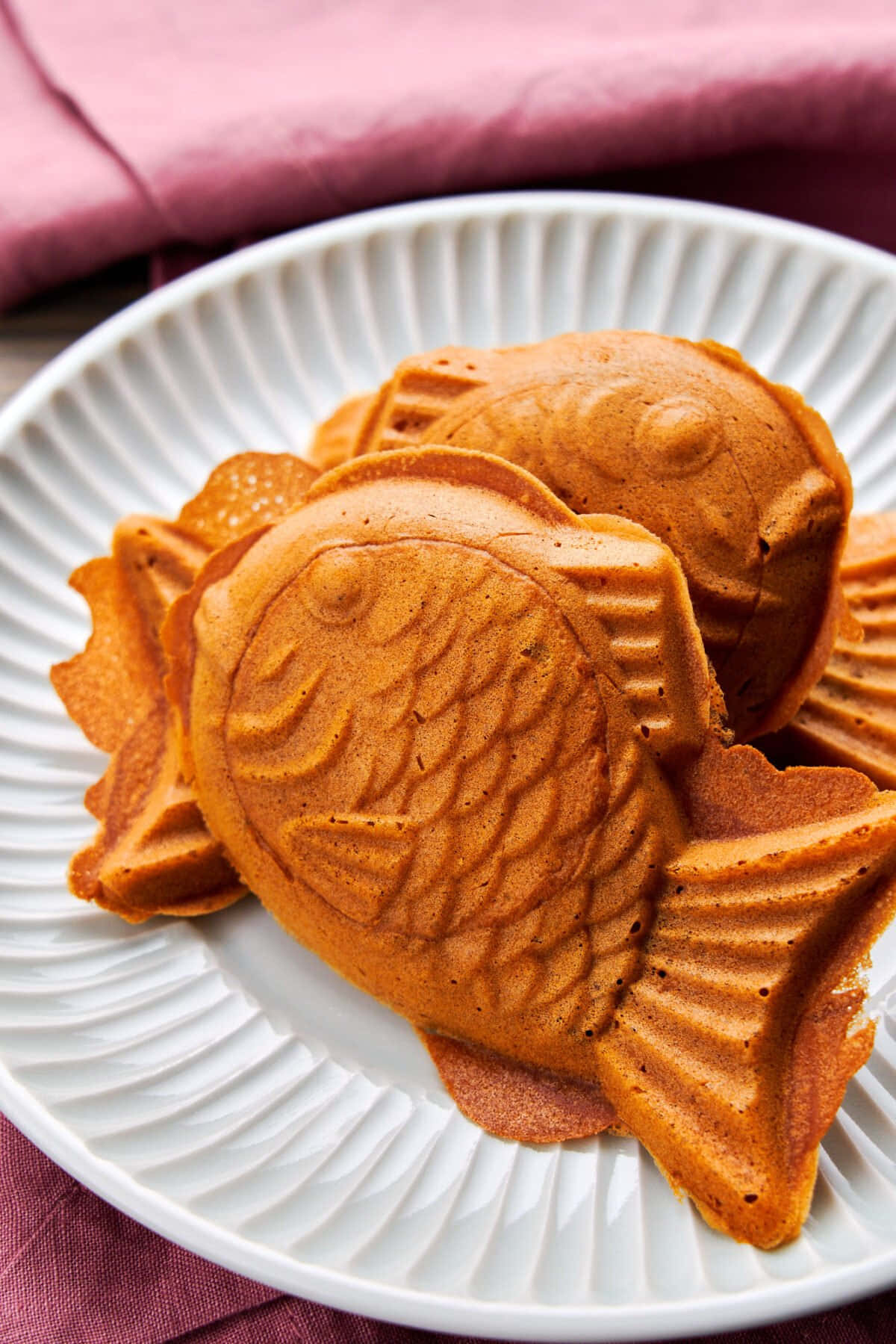 Japanese Sweets 桌布 壁紙 — 金棕色魚形鬆餅，帶有細緻鱗片紋理，堆疊在白色條紋盤上，襯以柔軟粉紅色布料。