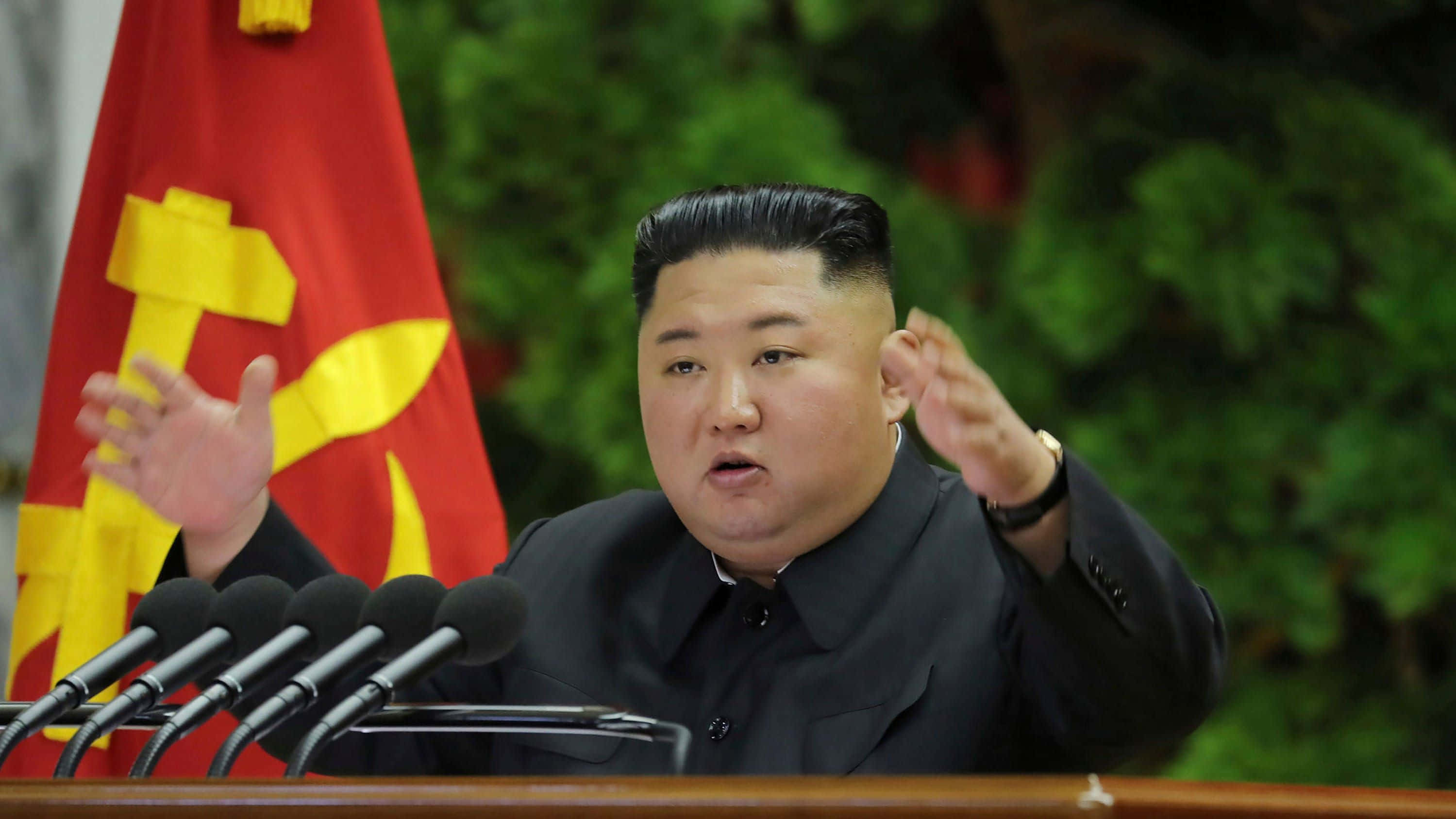 Kim Jong Un 桌布 壁紙 — 金正恩在帶有黃色鎚子與鐮刀的紅旗前，於設有麥克風的講台上強烈地做手勢發言。