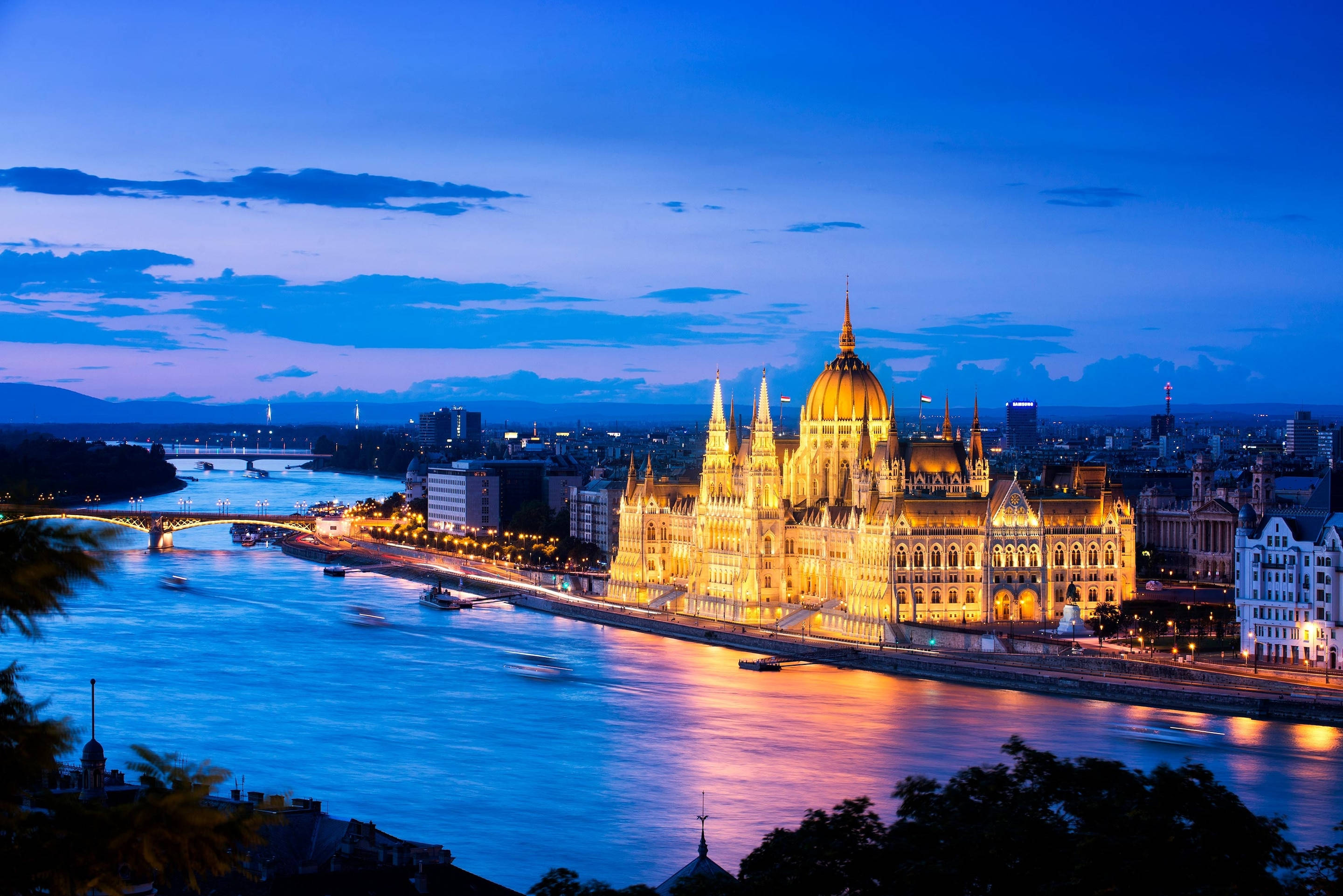 budapest 桌布 壁紙 — 金碧輝煌的匈牙利國會大廈在黃昏的天空下，在多瑙河畔溫暖地發光，城市燈光倒映在水面上。