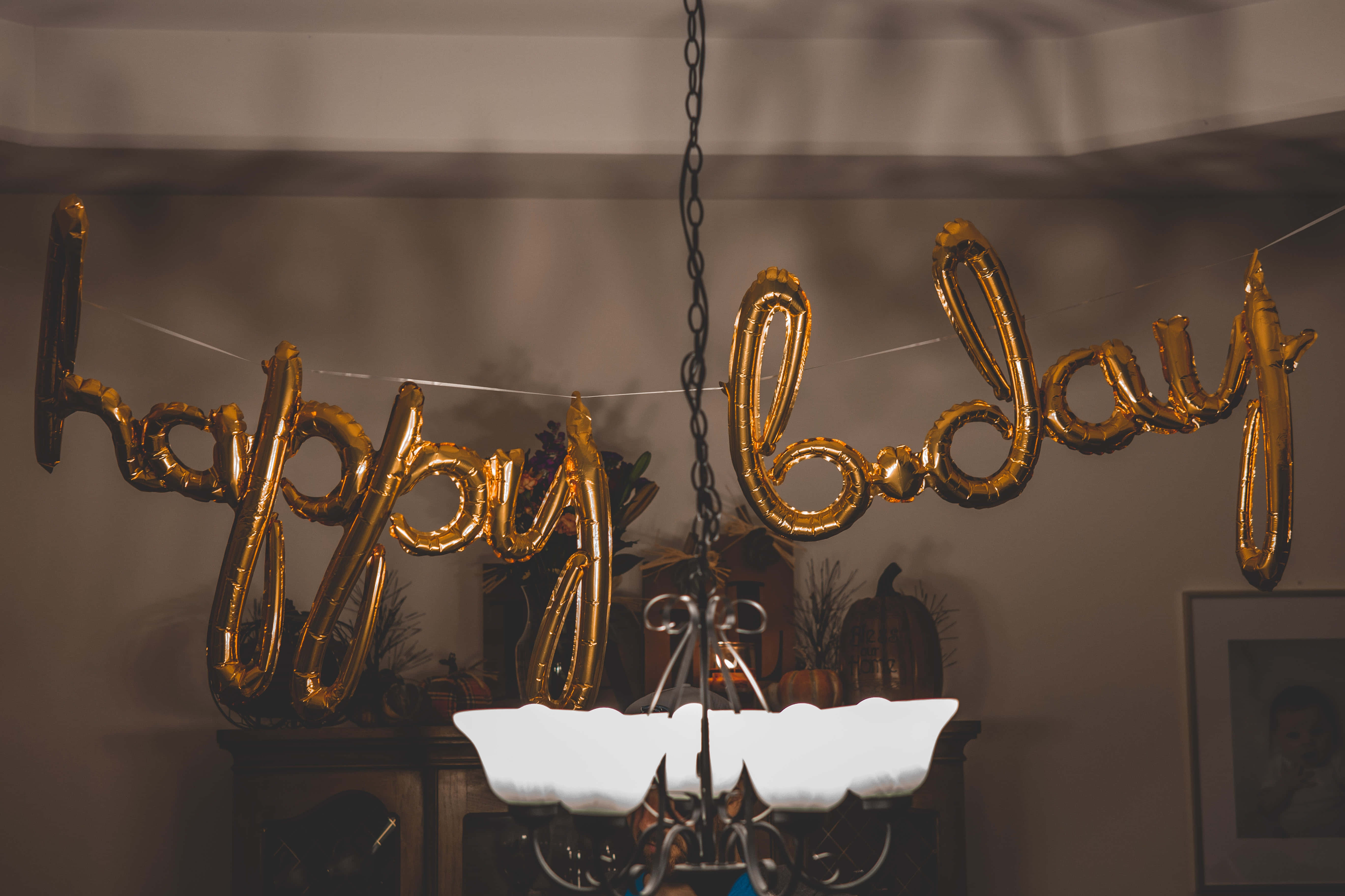 Cute Birthday 桌布 壁紙 — 金色 'happy birthday' 氣球懸掛在點亮的吊燈上方，營造出溫暖的室內慶祝場景。