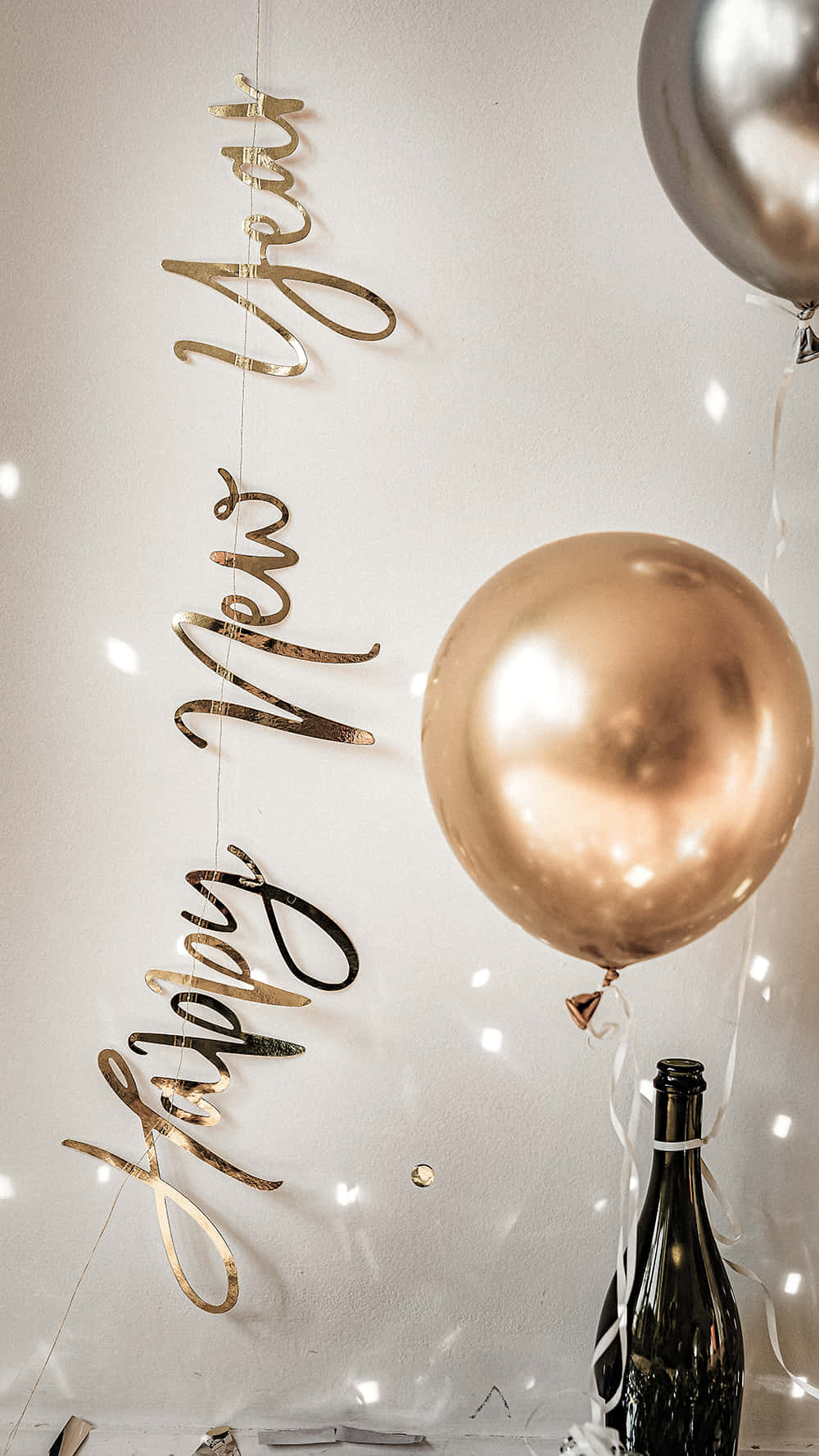桌布 壁紙 — 金色 'Happy New Year' 字母掛在玫瑰金氣球和香檳瓶旁邊，背景是柔和照明的白色牆壁。