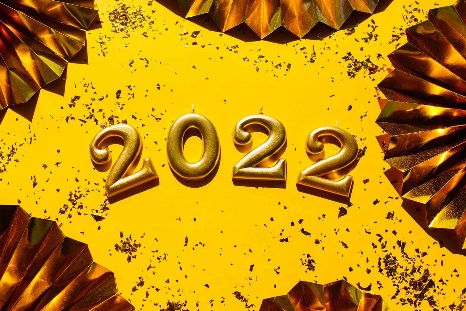 新年 桌布 壁紙 — 金色2022蠟燭放在明亮的黃色背景上，周圍是金屬扇裝飾和五彩紙屑。
