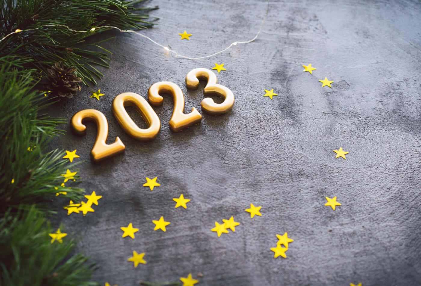 新年 桌布 壁紙 — 金色2023數字和黃色星星放在深色紋理表面上，旁邊有松樹枝和松果。