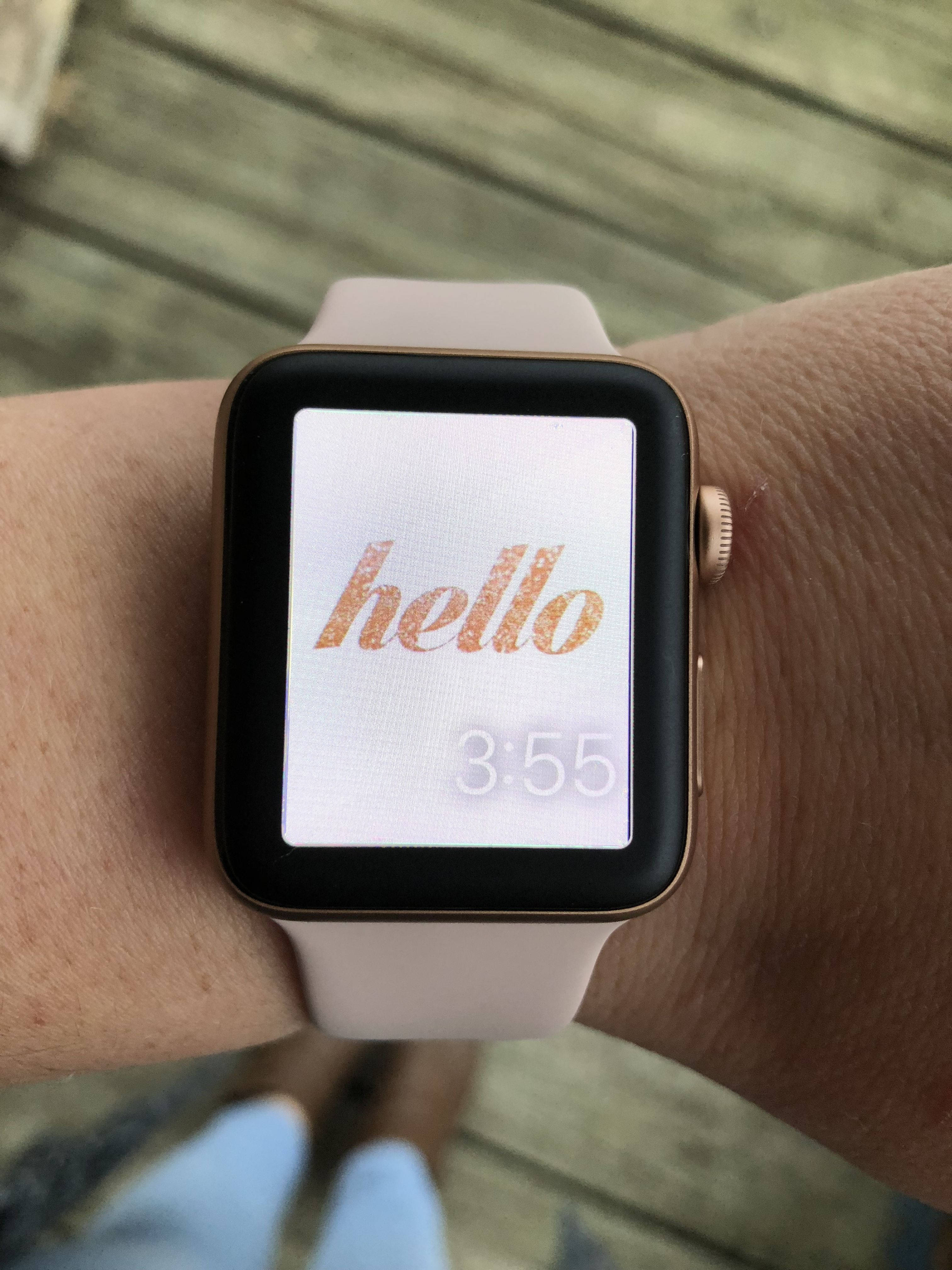Apple Watch 動態 桌布 壁紙 — 金色Apple Watch的特寫，搭配淺粉紅色錶帶，在木質甲板背景上以玫瑰金色草書顯示單詞hello和時間3:55。
