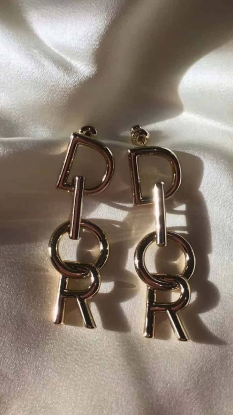 Dior 動態 桌布 壁紙 — 金色Dior標誌耳環放在柔軟的白色緞面上，定向燈光投射出戲劇性的陰影。