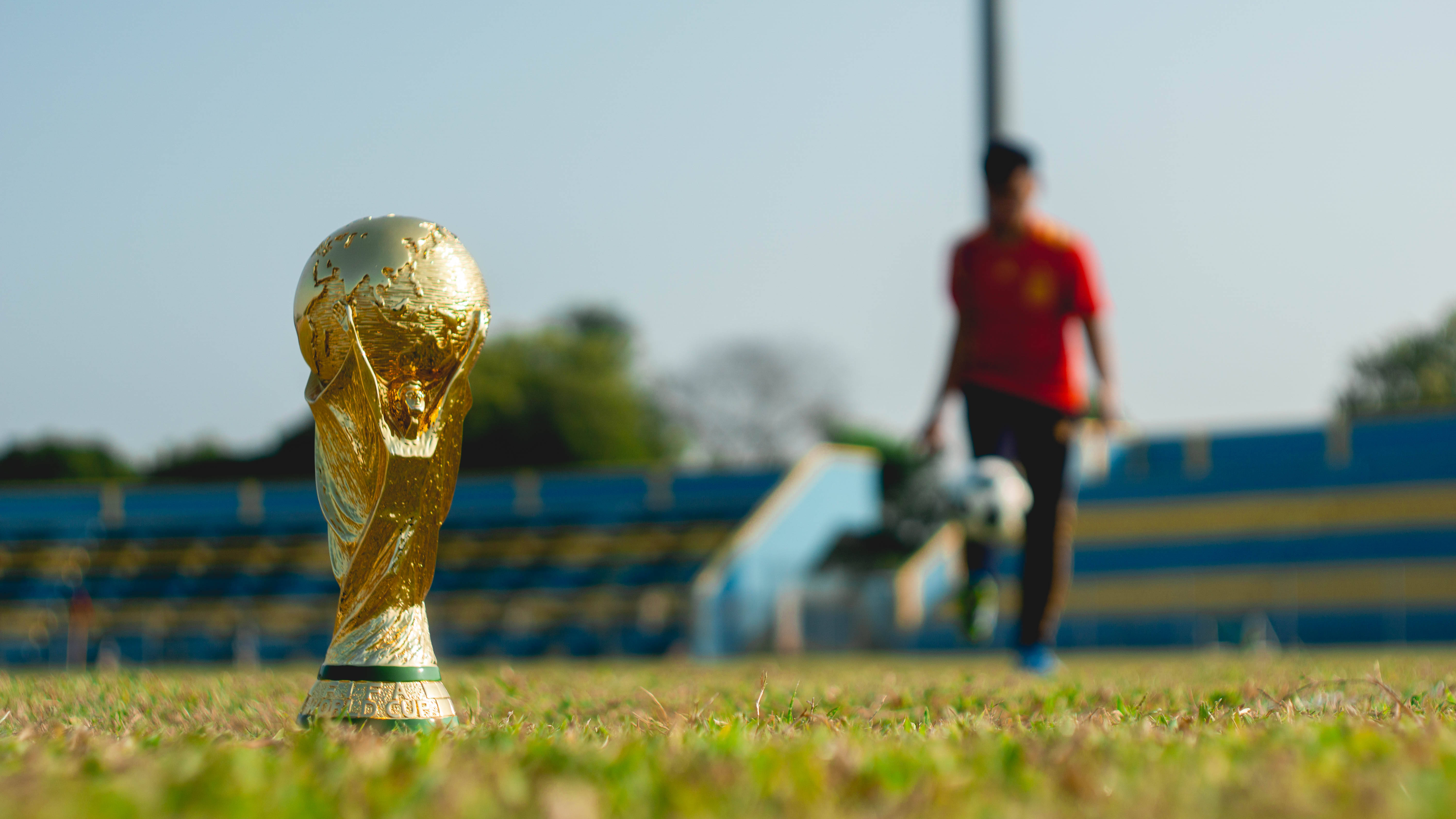 World 桌布 壁紙 — 金色FIFA世界盃獎盃在草地上閃耀，背景中一個模糊的紅色足球員拿著球走過。