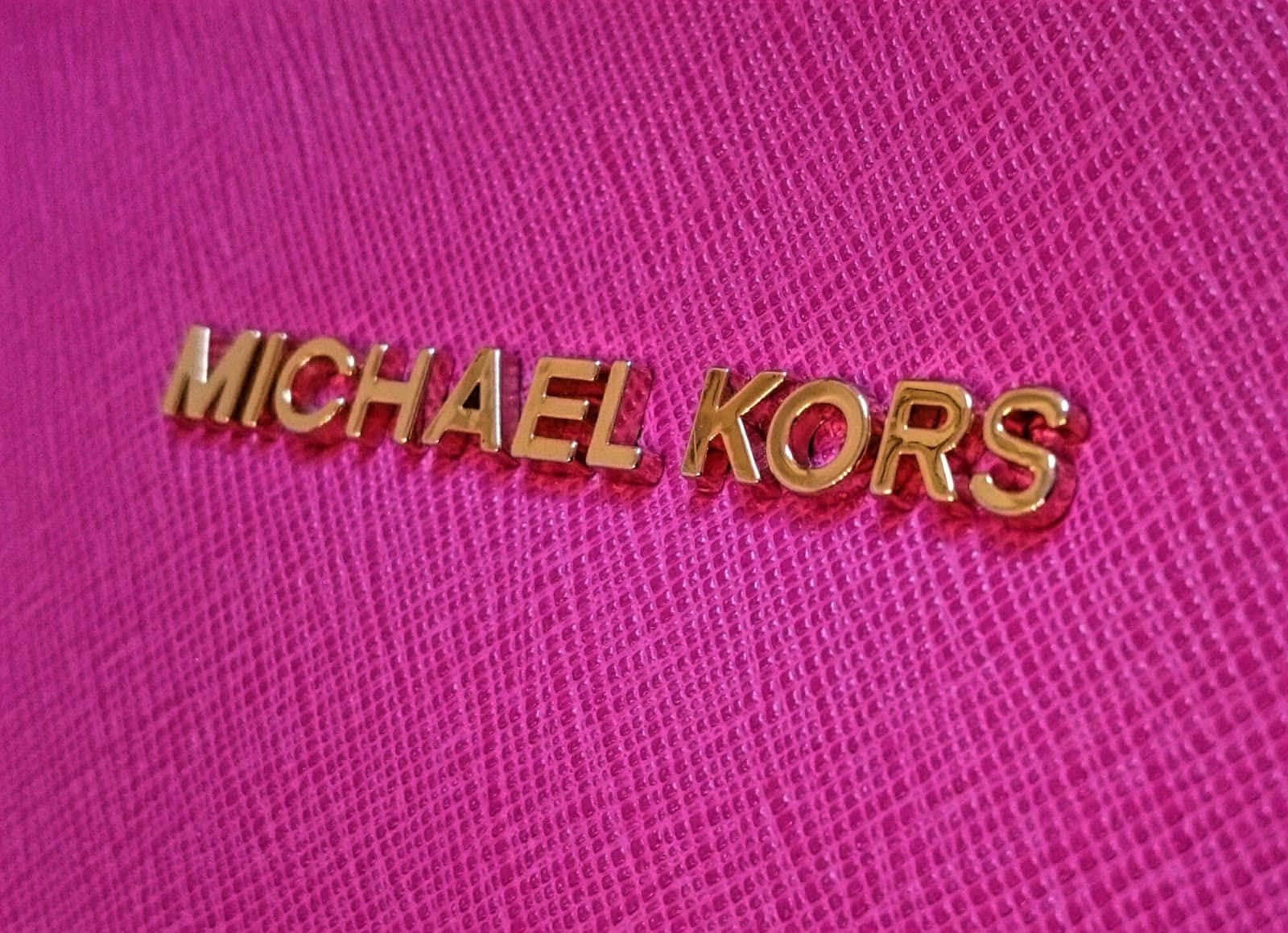 麥可·寇斯 桌布 壁紙 — 金色Michael Kors標誌在紋理熱粉紅色布料上的特寫，對焦銳利，陰影柔和。