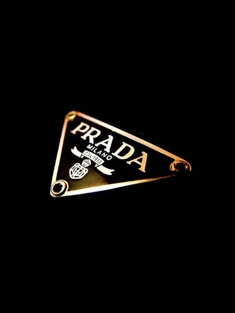 Prada 桌布 壁紙 — 金色Prada三角形標誌胸針在戲劇性燈光下於純黑背景上閃耀。