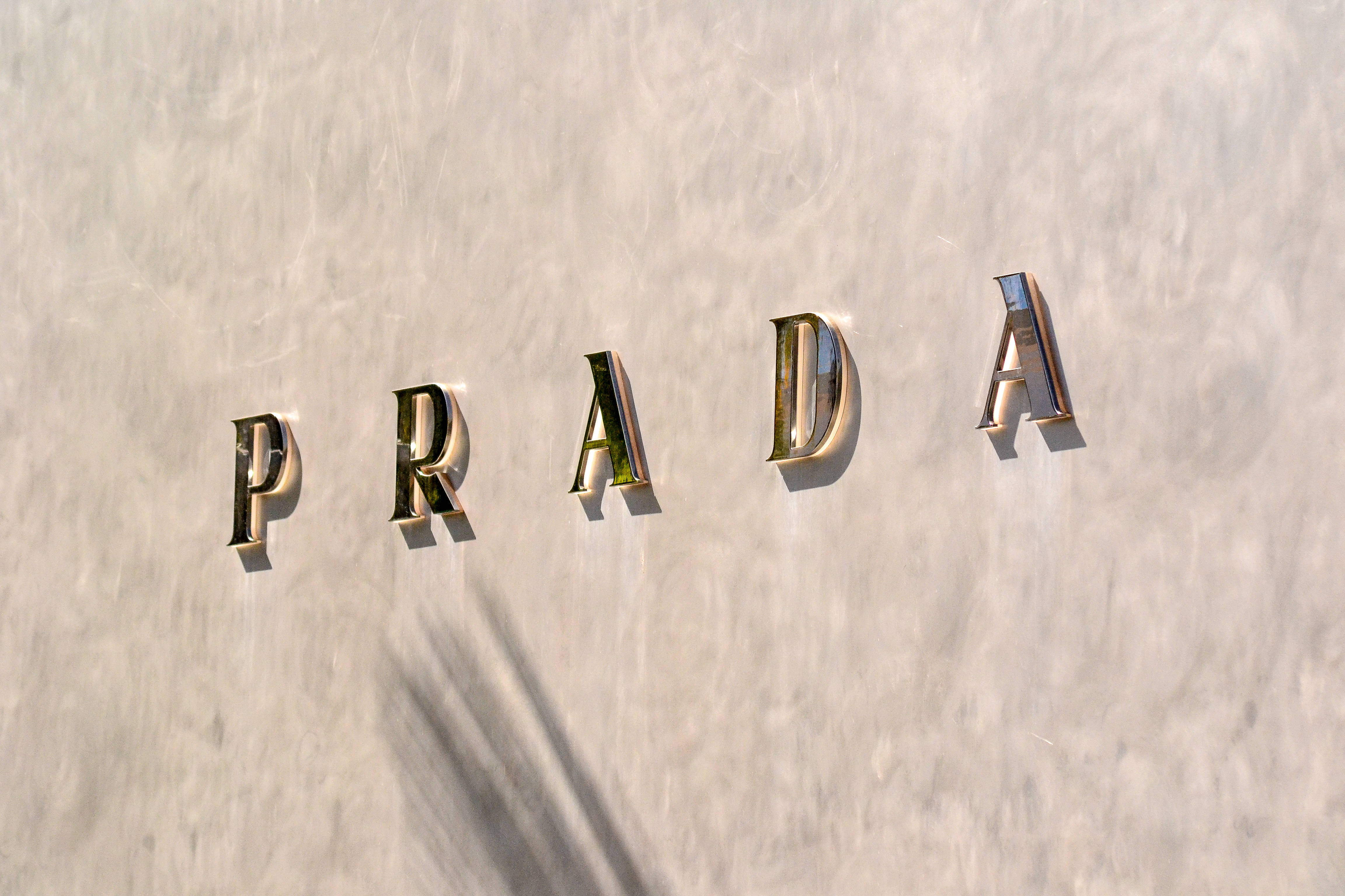Prada 桌布 壁紙 — 金色Prada字母安裝在帶有紋理的米白色牆壁上，表面投射出棕櫚樹的影子。