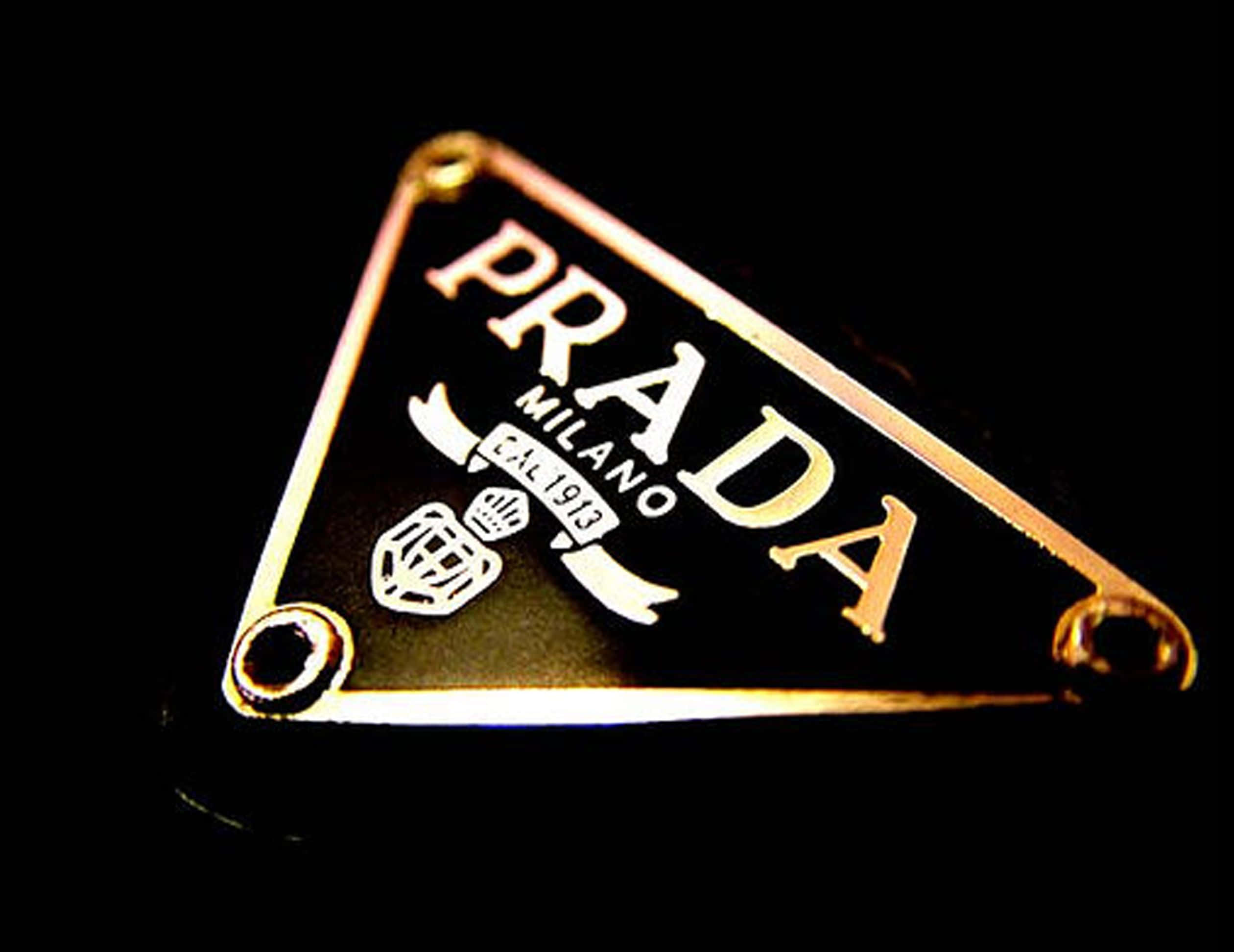 Prada 桌布 壁紙 — 金色三角形Prada標誌牌，上面有白色蝕刻的「MILANO DAL 1913」，背景為黑色。