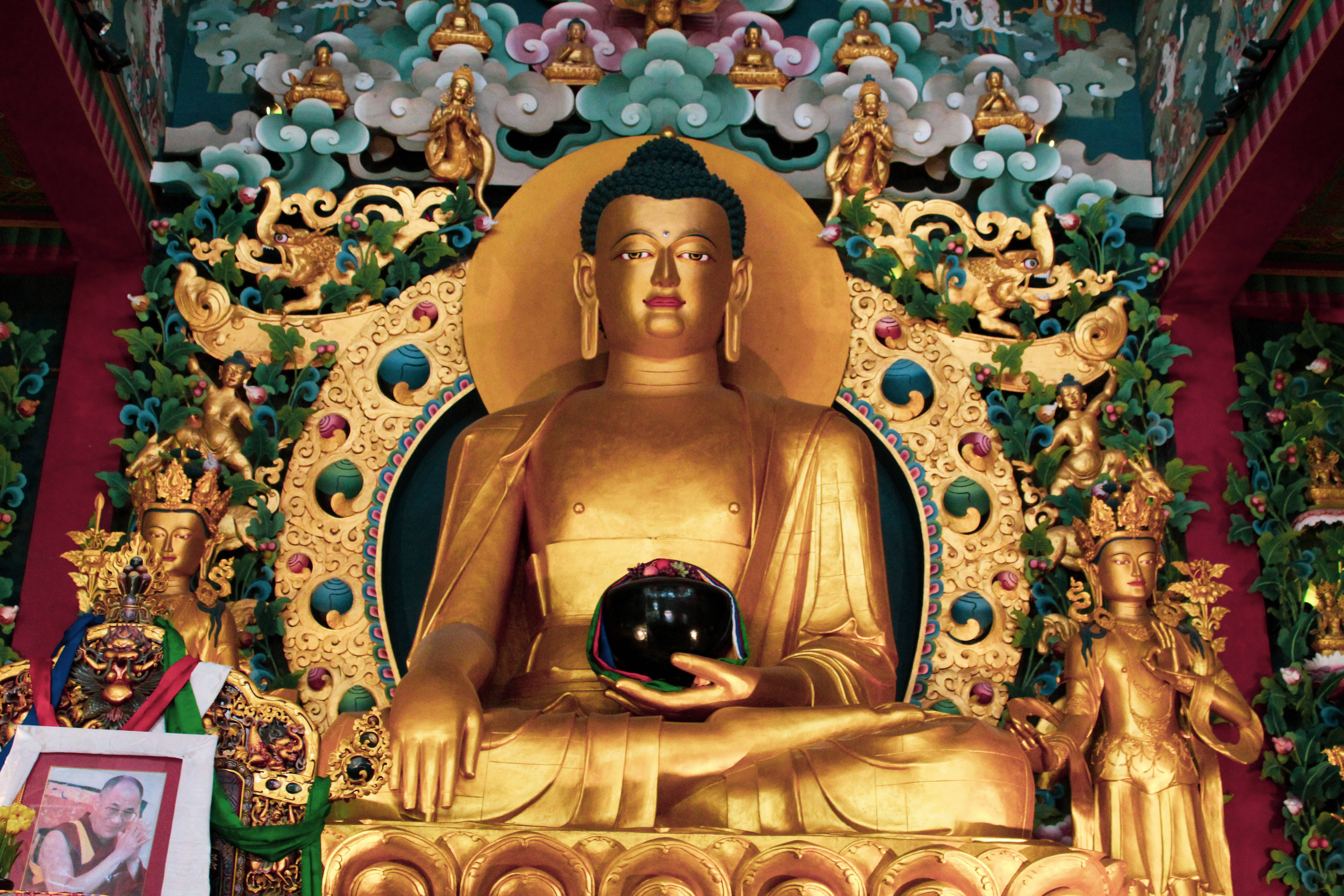 Gautam Buddha 桌布 壁紙 — 金色佛像以冥想姿勢坐著，手持黑色球體，周圍環繞著華麗的神像和色彩繽紛的花卉裝飾。