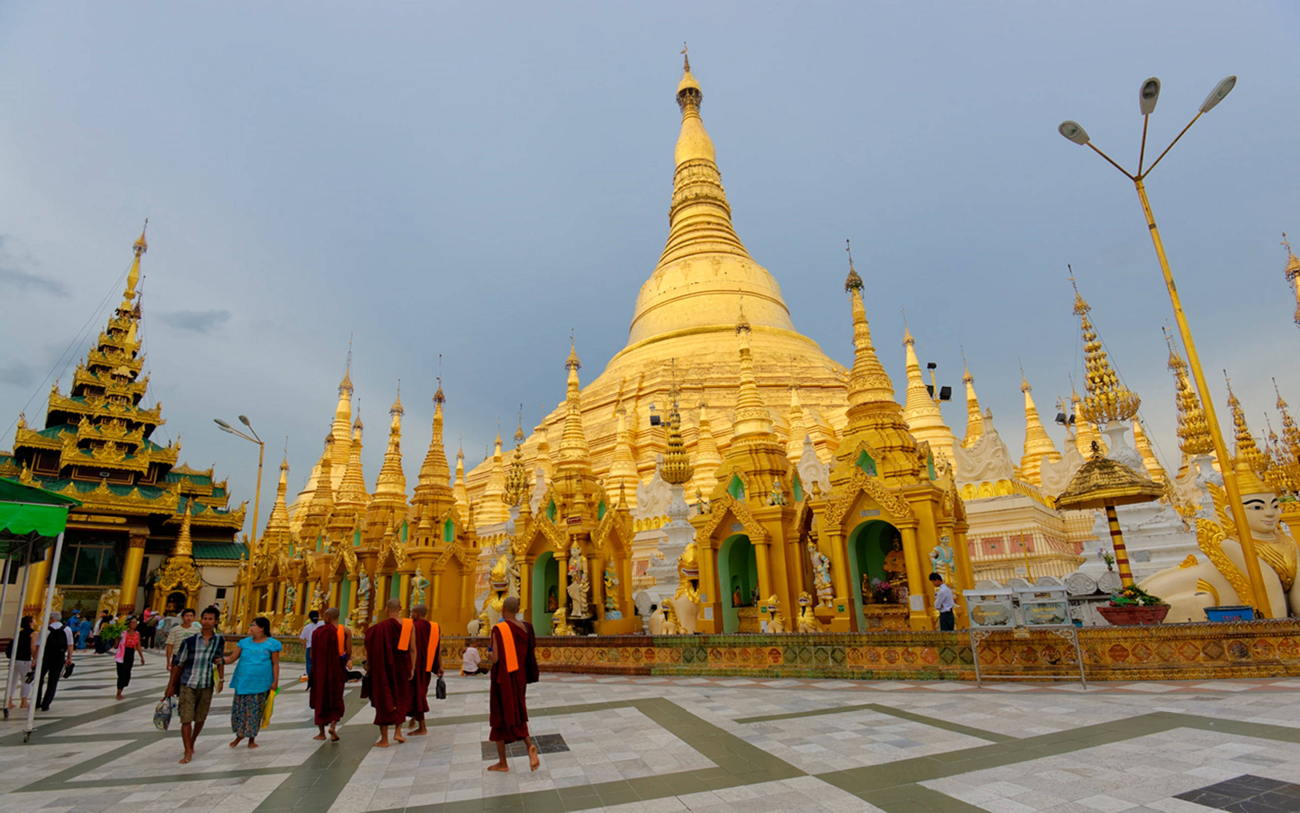 Yangon 桌布 壁紙 — 金色佛塔在柔和天空下閃耀，僧侶身穿栗色長袍走過瓷磚廣場。