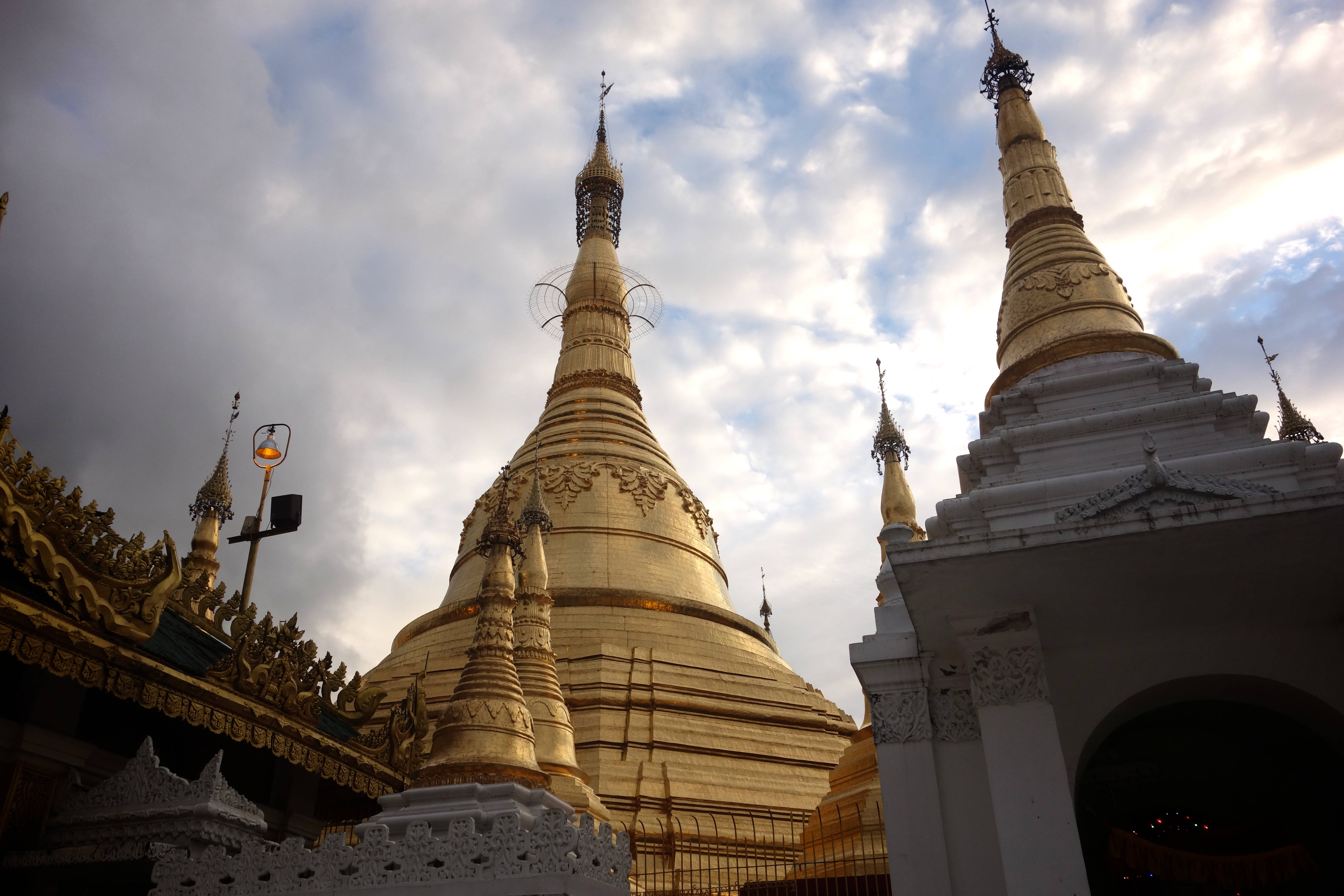 Yangon 桌布 壁紙 — 金色佛塔在陰雲密佈的天空下巍然聳立，精緻的尖頂和華麗的白色建築構成了畫面的框架。