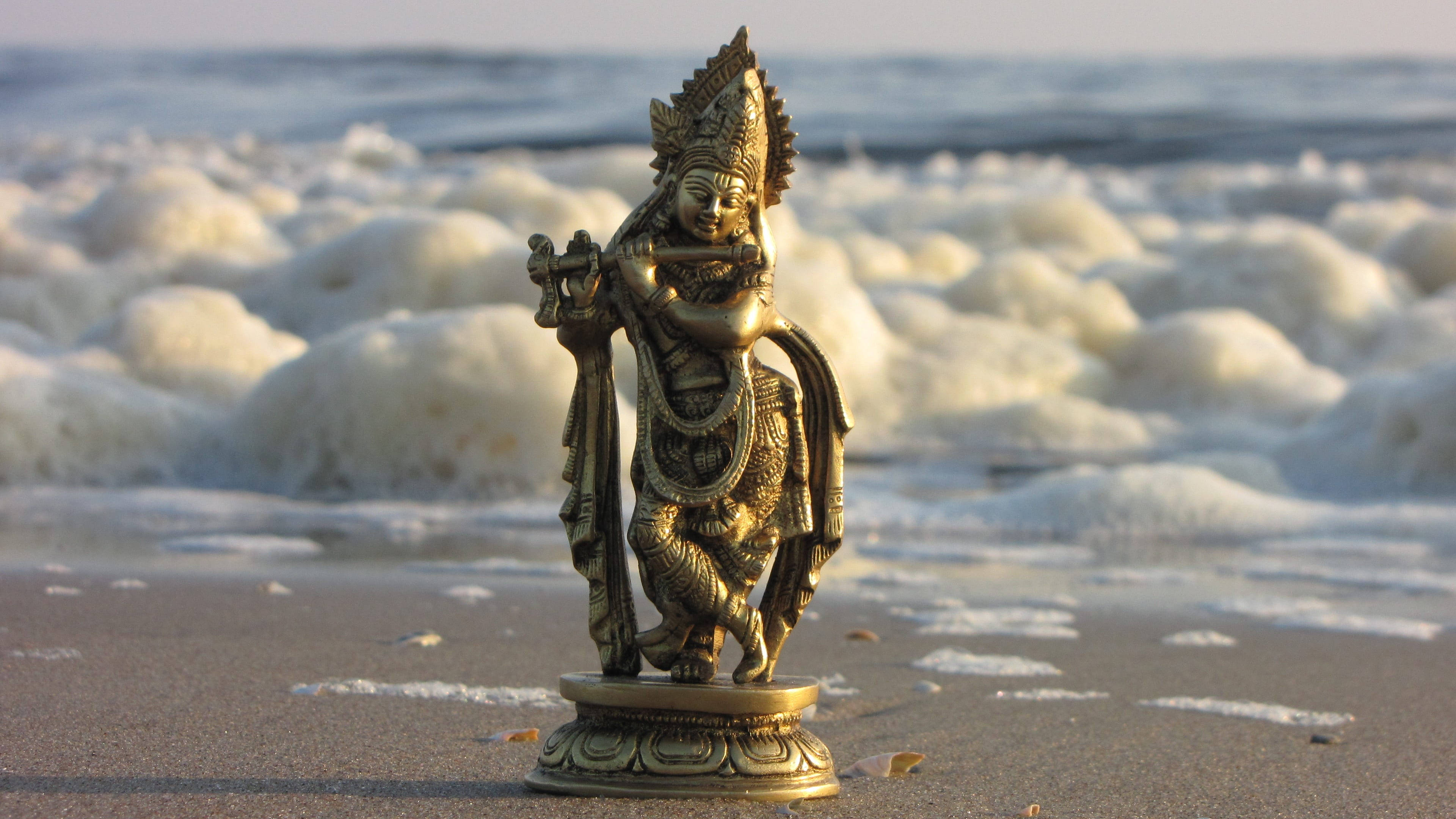 Shri Krishna 桌布 壁紙 — 金色克里須那神像在沙灘上吹奏長笛，背後海浪拍打，柔和陽光灑落。